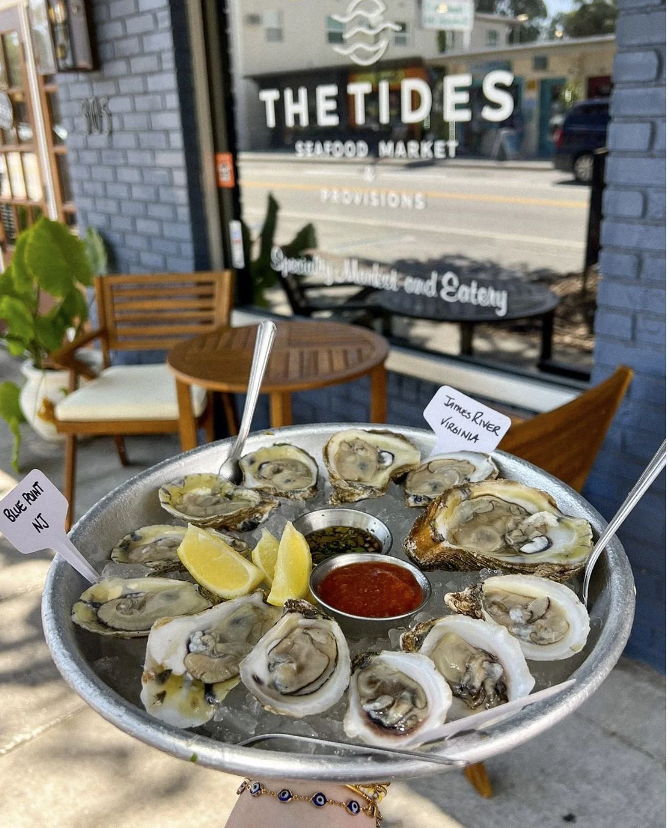A plate of fresh oysters on ice with lemon wedges, dipping sauces, and small tags indicating the oyster sources, held outside a seafood market called 'The Tides' with outdoor seating and a brick building in the background.