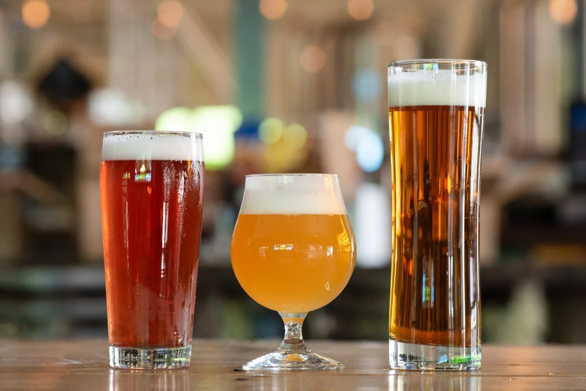 Three different types of beer in various glasses on a wooden bar, with a blurred background.