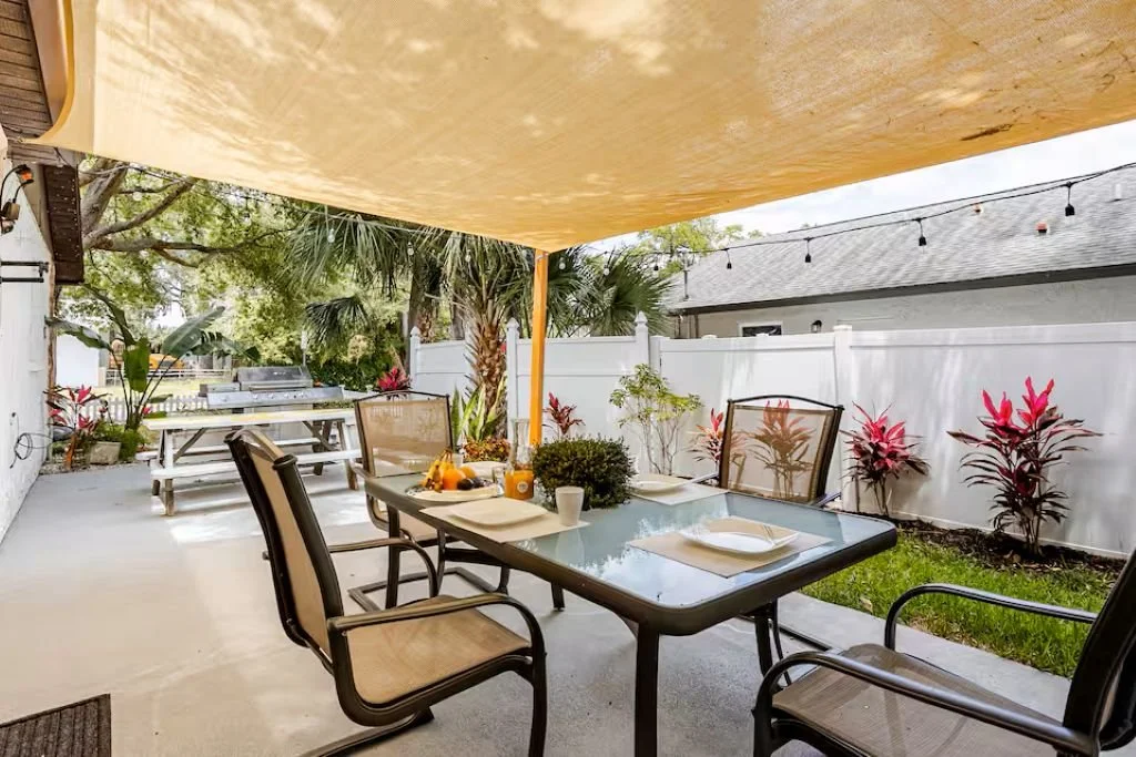Backyard patio with a dining table, chairs, and plants, enclosed by a white fence and a canopy overhead.