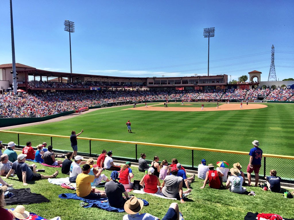 View of a baseball game with spectators seated on the grass and in the stands, players on the field, and a clear blue sky.