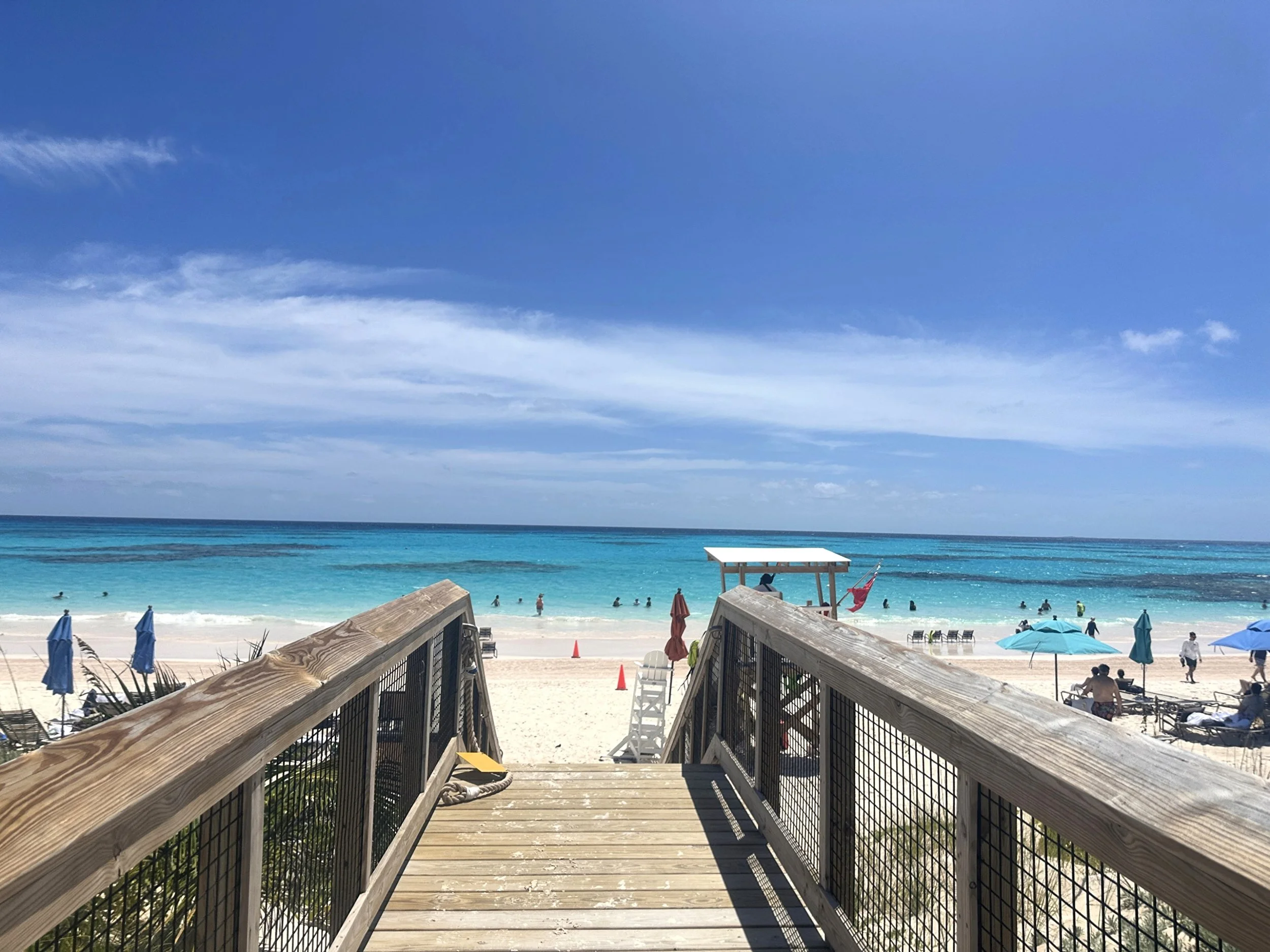 Wooden walkway leading to a beach with blue umbrellas, sunbathers, and ocean waves under a partly cloudy sky.