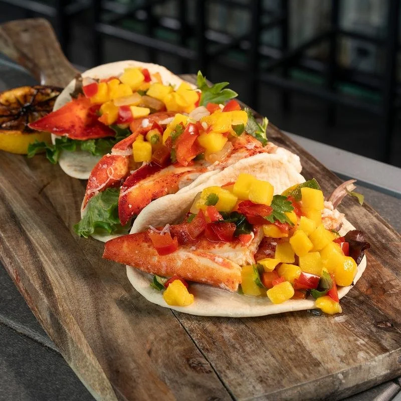 Three lobster tacos with diced mango, tomatoes, and cilantro on a wooden serving board.