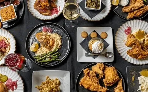 Variety of fried chicken, mashed potatoes with gravy, green beans, macaroni and cheese, and drinks on a black table with paper plates and bowls.