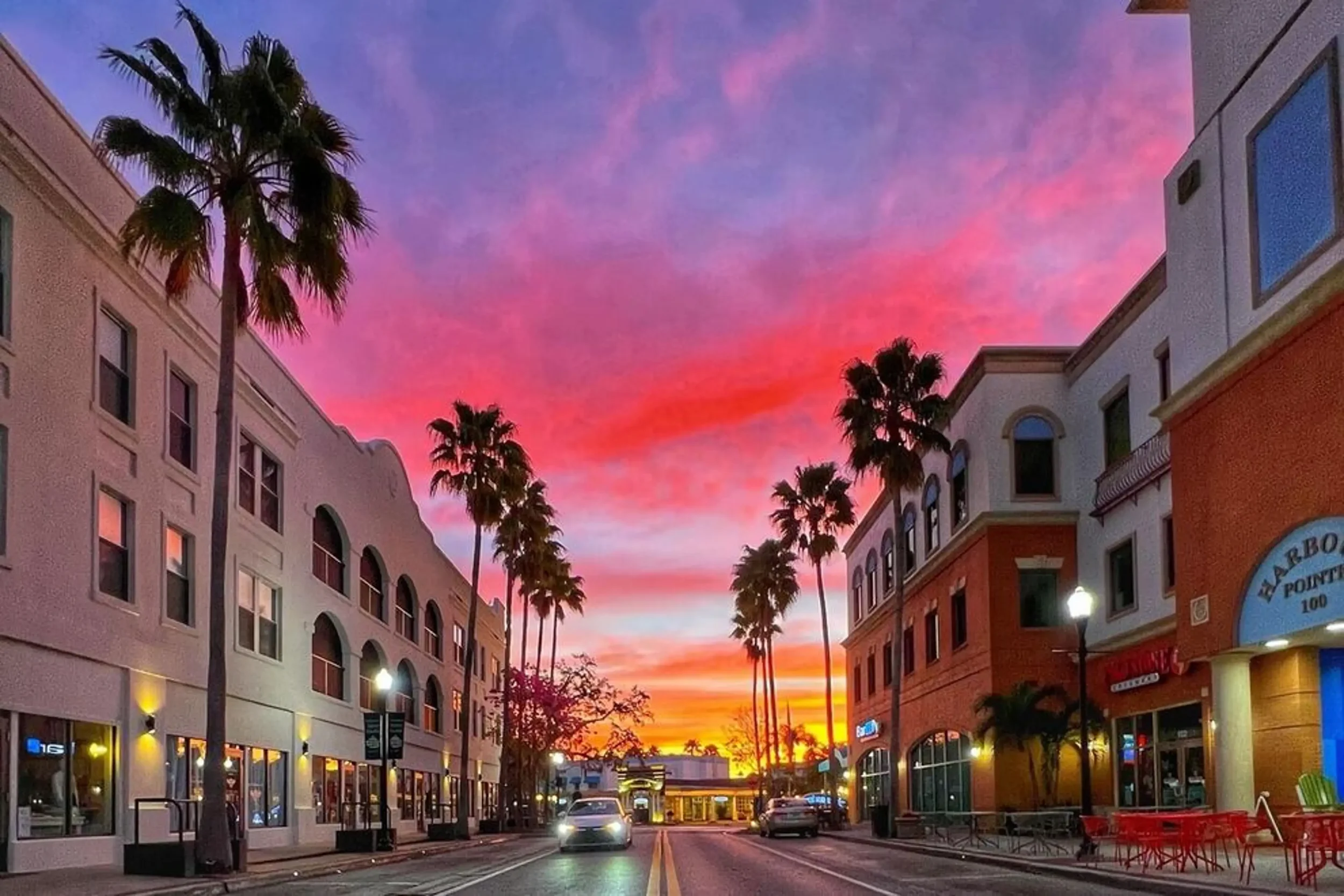 City street during sunset with colorful pink, purple, and orange sky, lined with palm trees and multi-story buildings, some with lit windows, and parked cars along the street.