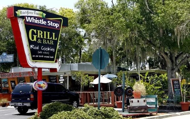 Sign for Whistle Stop Grill & Bar with a black SUV passing by, outdoor seating area with tables and umbrellas, trees, and a sidewalk in front.