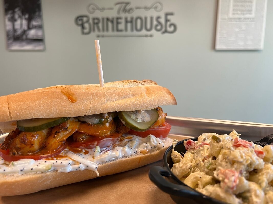 A sandwich with grilled chicken, pickles, tomato, and sauce in a hoagie roll, accompanied by a side of potato salad, on a tray at a restaurant called Brinehouse.