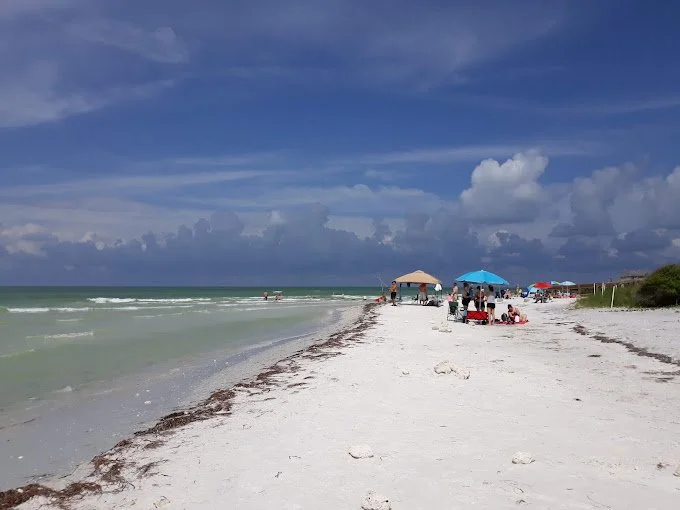 People relaxing under umbrellas on a sandy beach with ocean waves under a partly cloudy sky.