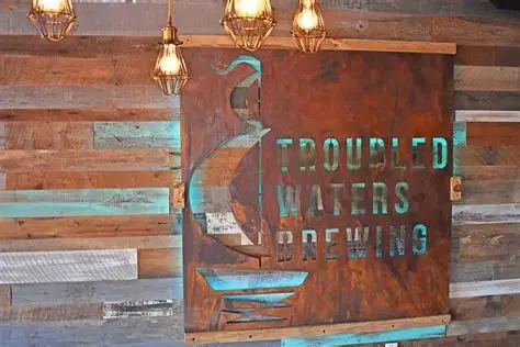 Rusty metal sign with cutout words 'TROUBLED WATERS BREWING' and a seahorse silhouette, mounted on a wooden wall with hanging Edison bulbs above.