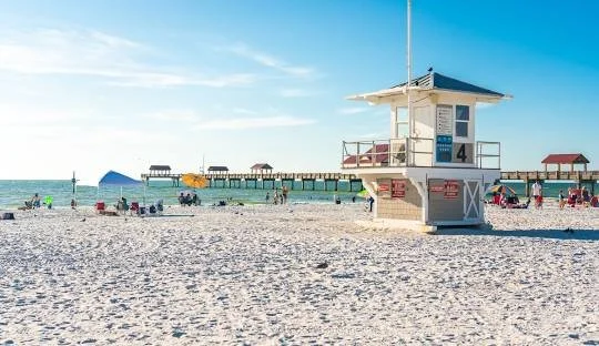 Empty beach with lifeguard tower, pier, and beach chairs under clear blue sky.