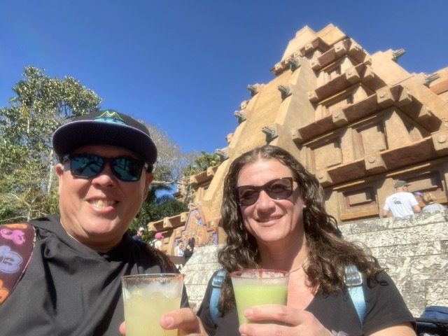 Two people smiling and holding green beverages in front of a large, pyramid-shaped building resembling a Mayan pyramid at an amusement park.