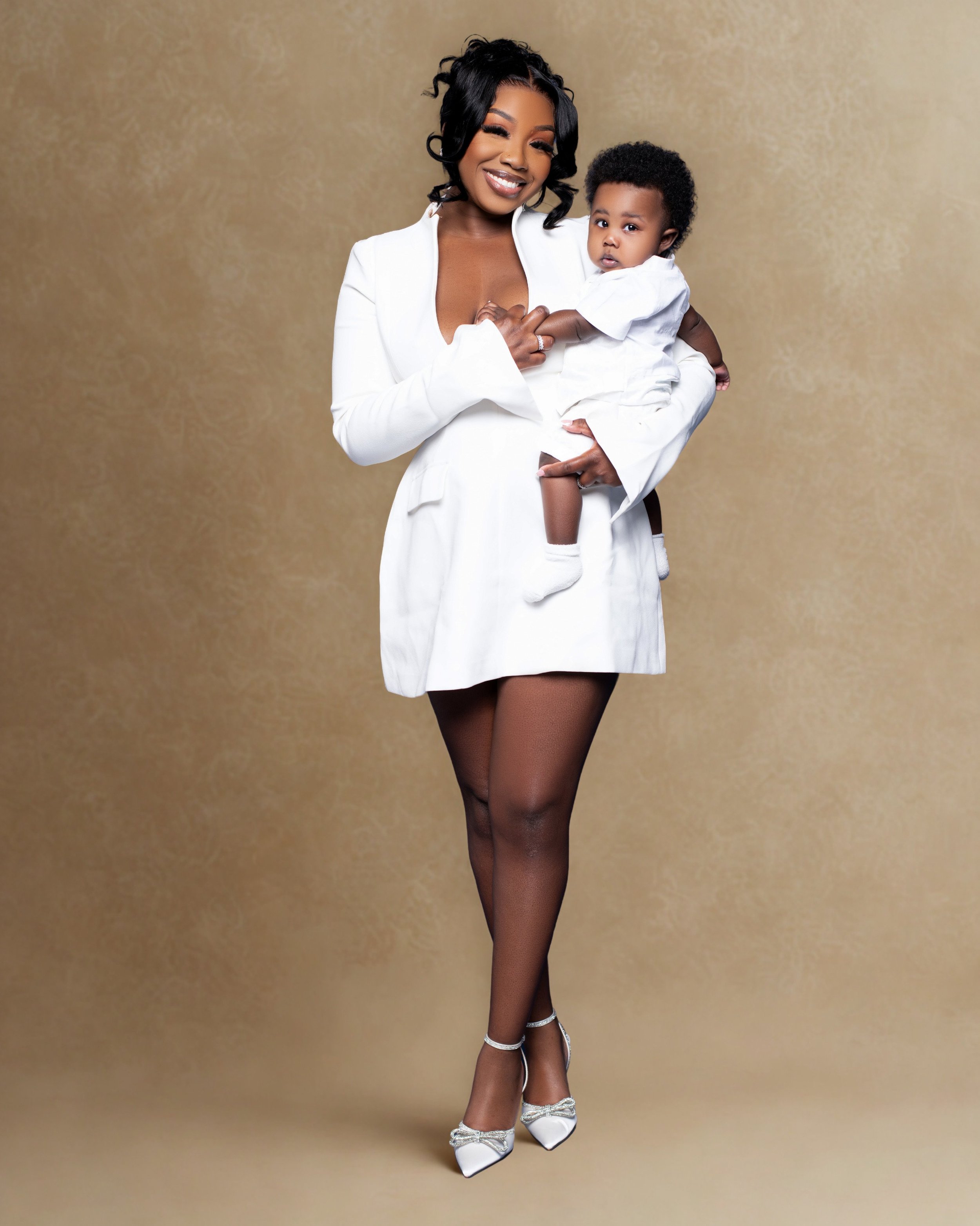 A woman dressed in a white blazer and thigh-high fishnet stockings holding a young child in a white shirt against a beige background.