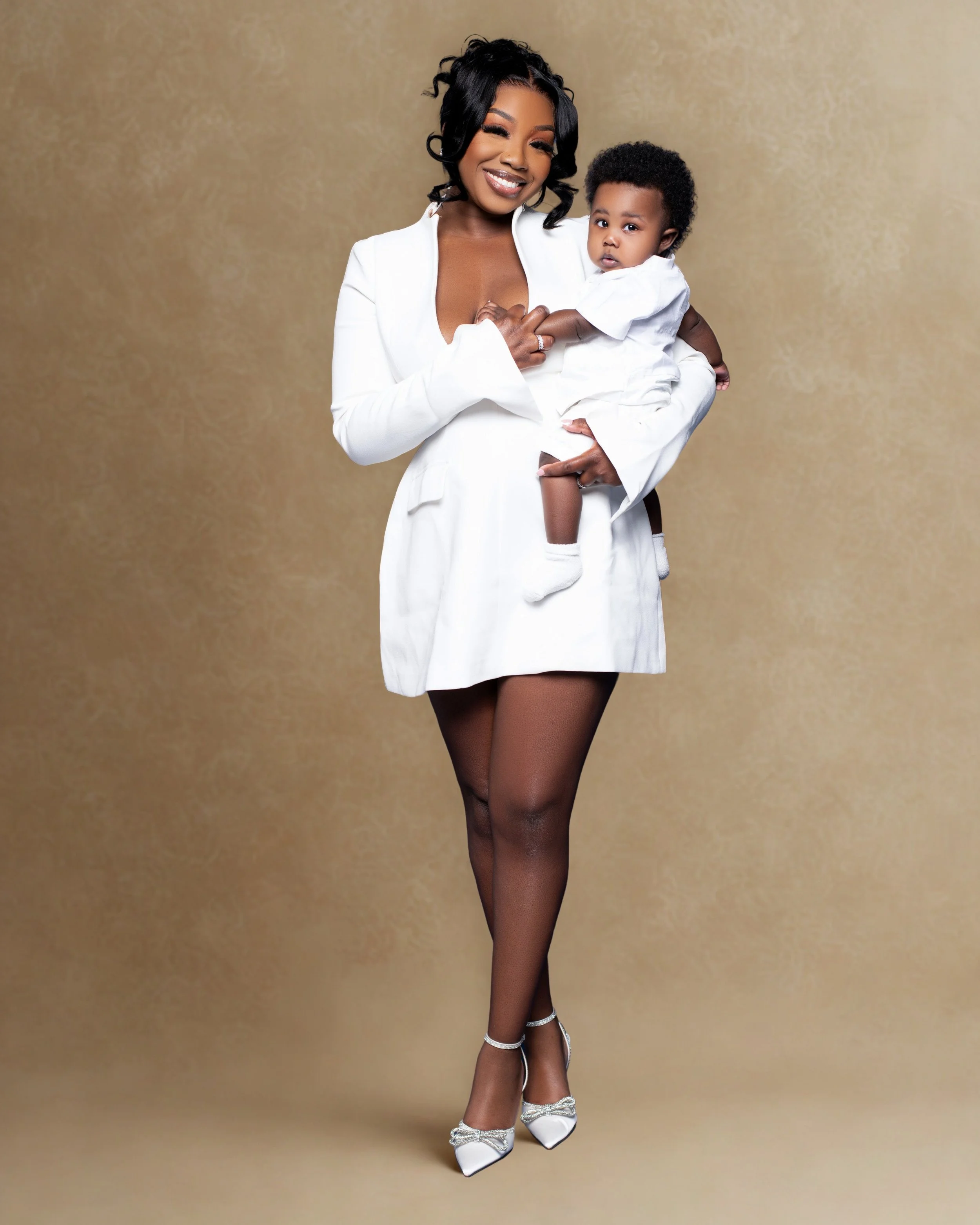 A woman with dark, curly hair smiling and holding a young child with short curly hair. The woman is dressed in a white blazer and black stockings with white high heels, standing against a beige textured background.