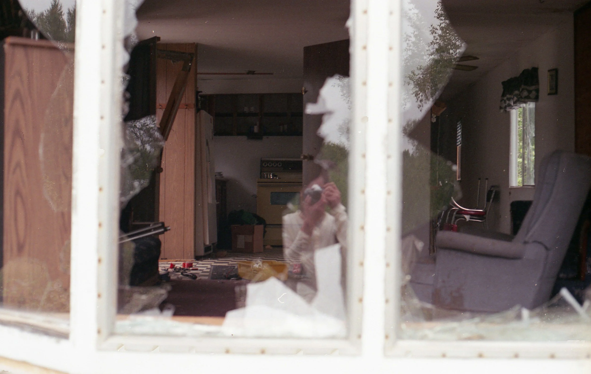 View of a messy living room through a broken window, with furniture, a checkered rug, and scattered objects visible inside.