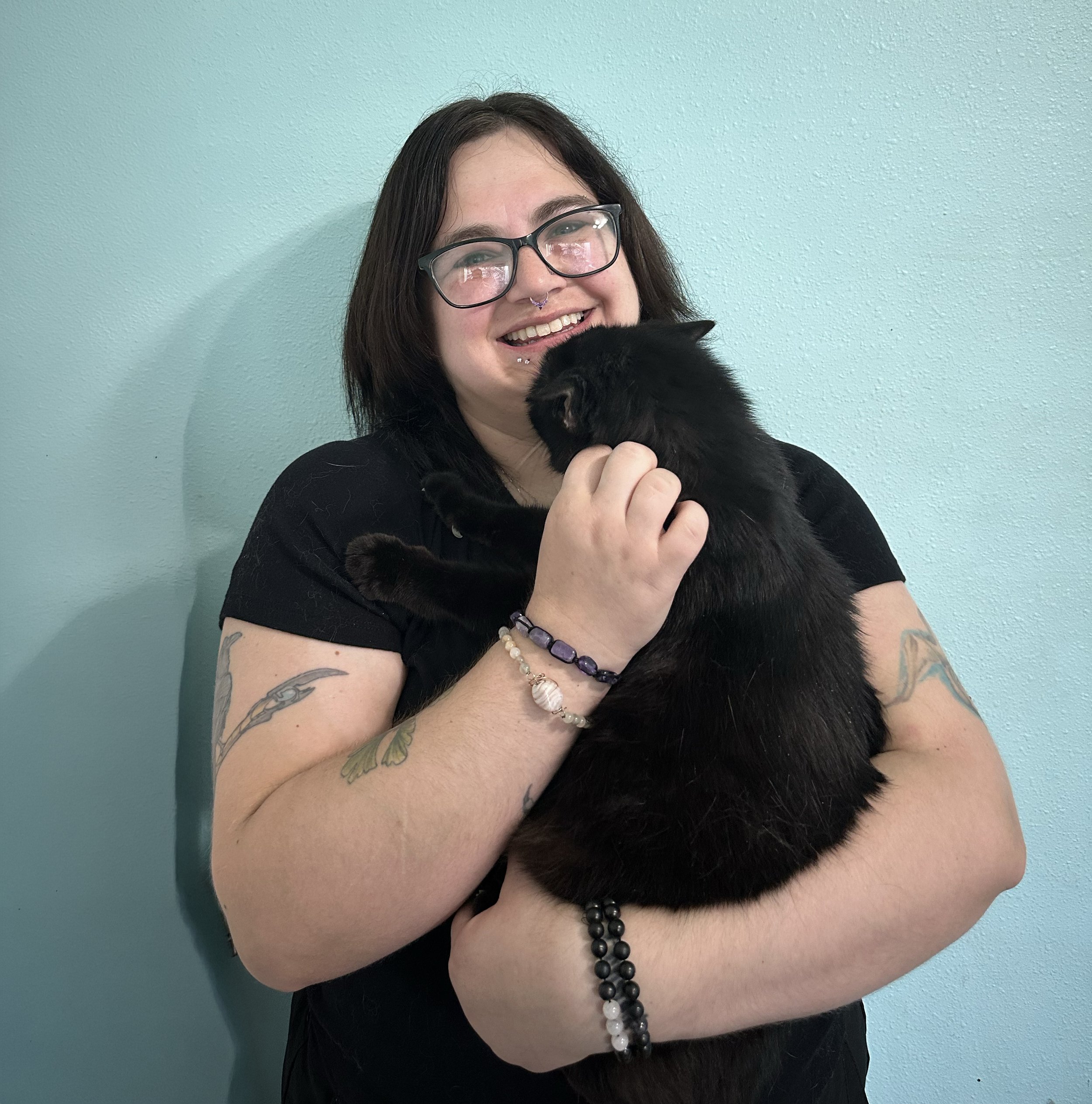 A woman with shoulder-length dark hair and glasses holding a black cat against a teal wall.