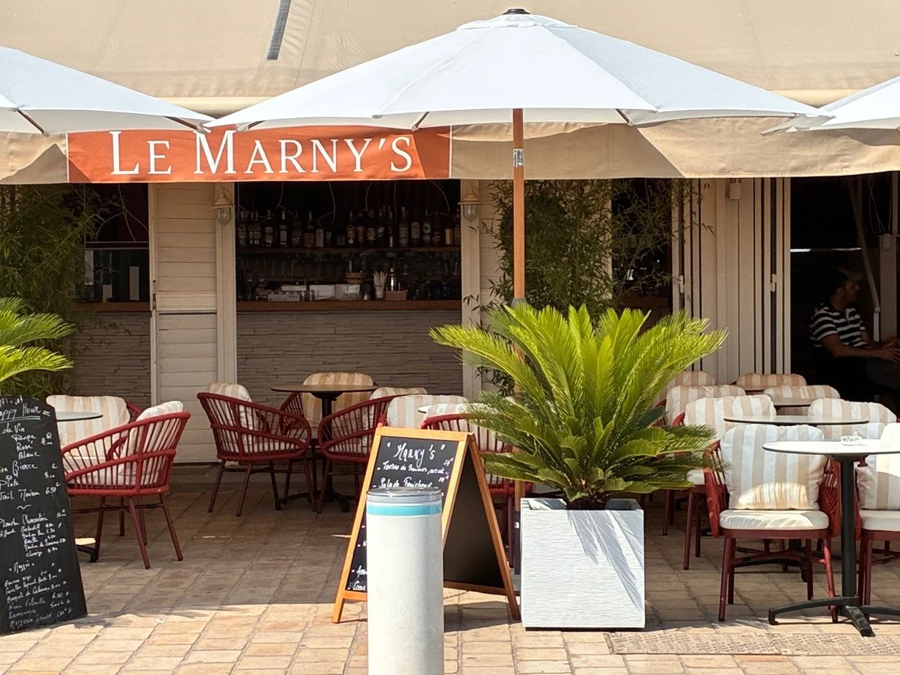 Terrasse d'un café nommé 'Le Marny's' avec des parasols blancs, des chaises rouges et des coussins beige et blanc, une grande plante verte en pot au centre, et un tableau noir avec le menu, en arrière-plan un bar et une personne assise.