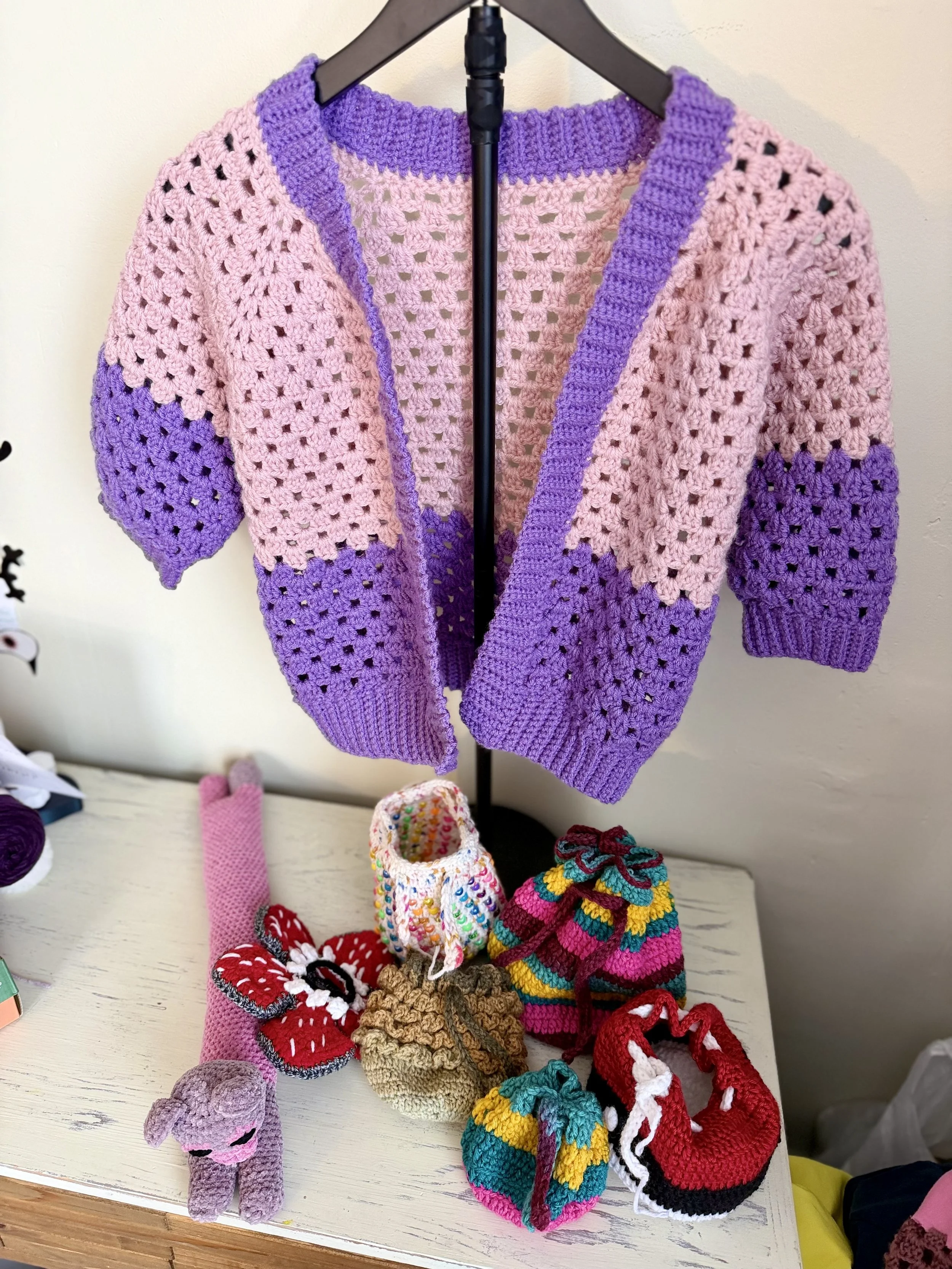 Display of various colorful handmade crocheted items, including slippers, hats, and stuffed animals, arranged on a table and hanging on a rack.