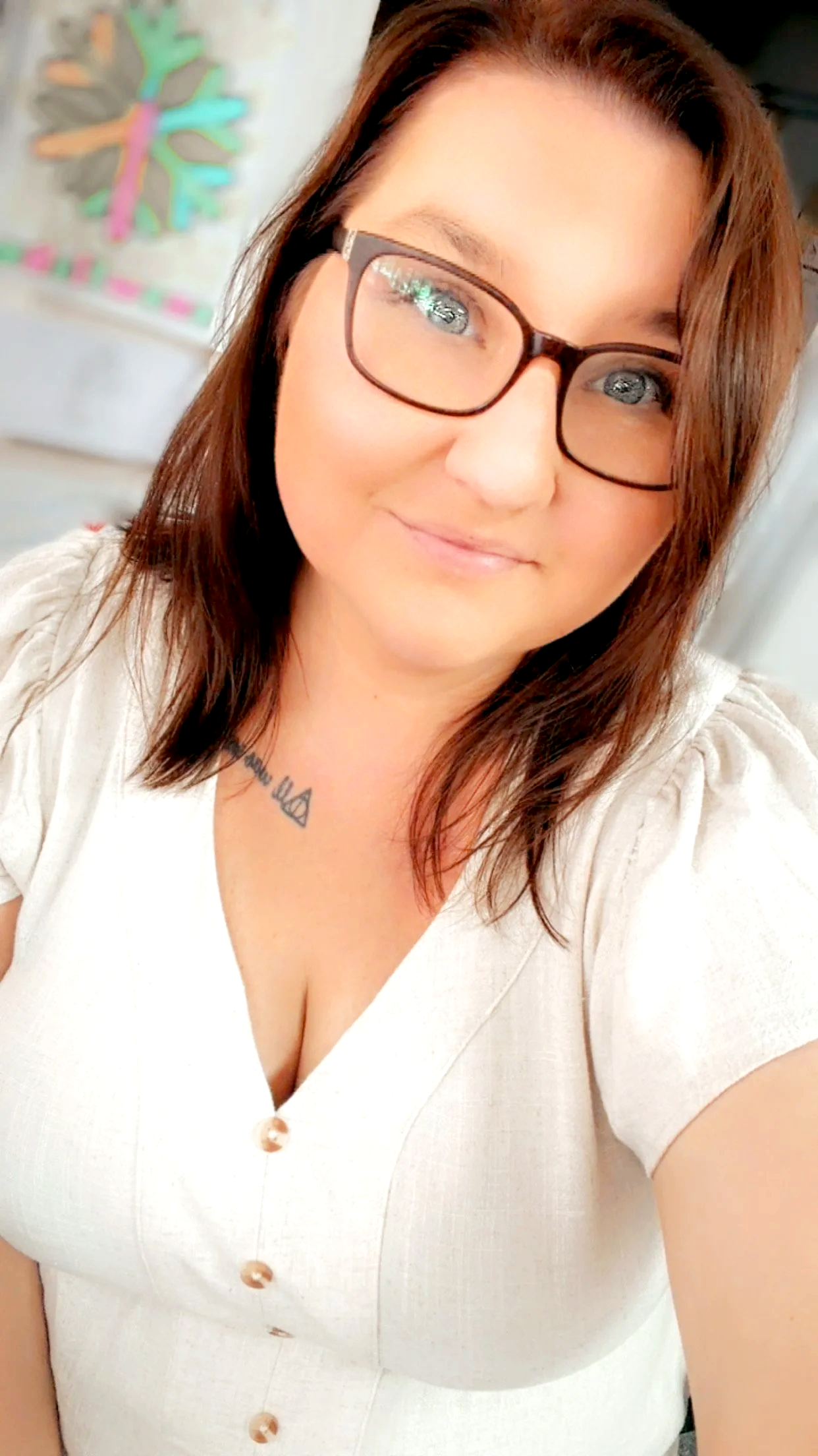 A woman with reddish-brown hair wearing glasses and a white top, smiling at the camera. There is a colorful art piece with a tree design in the background.