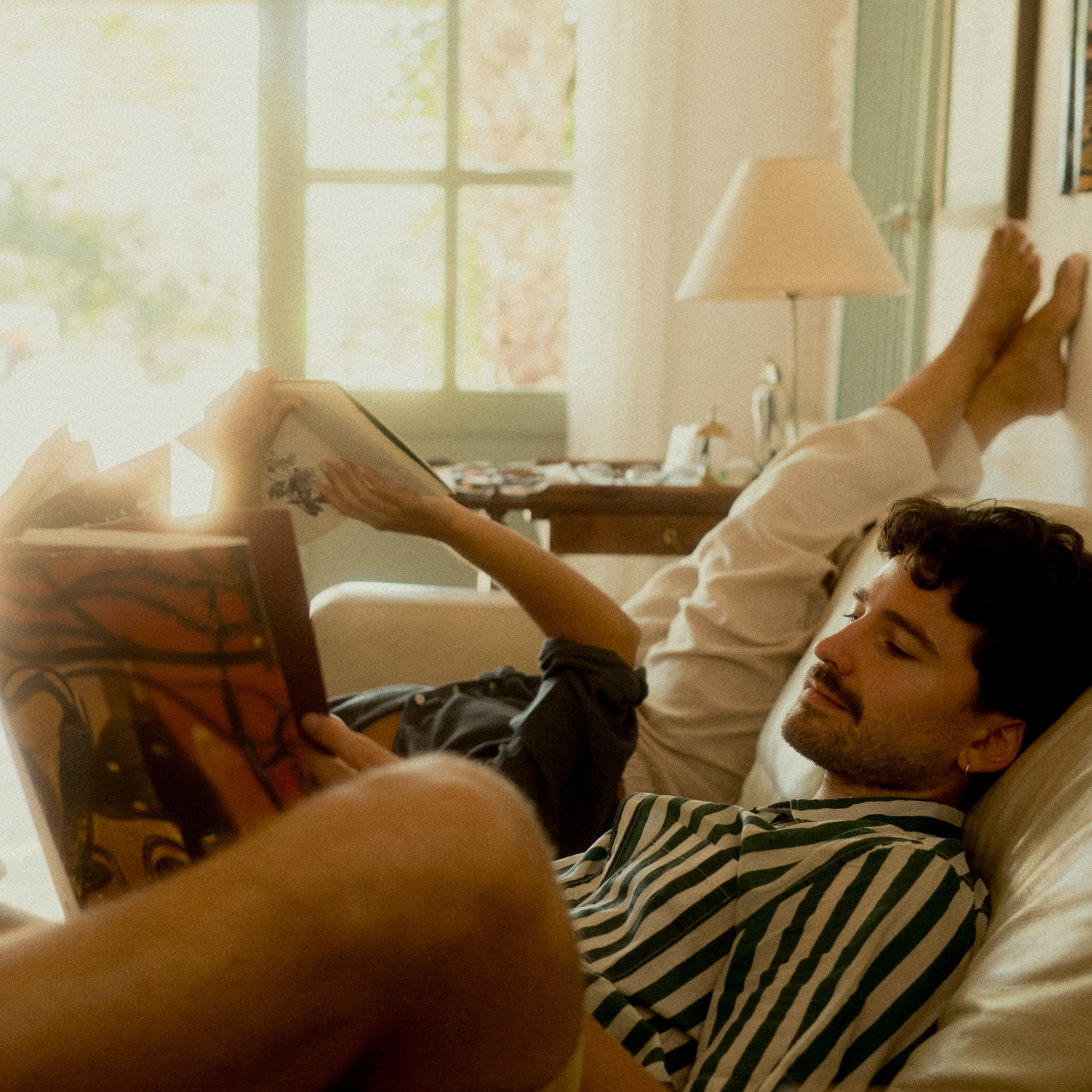 A person is lying on a bed, reading a book, with another person's feet visible in the background near a window with natural light.