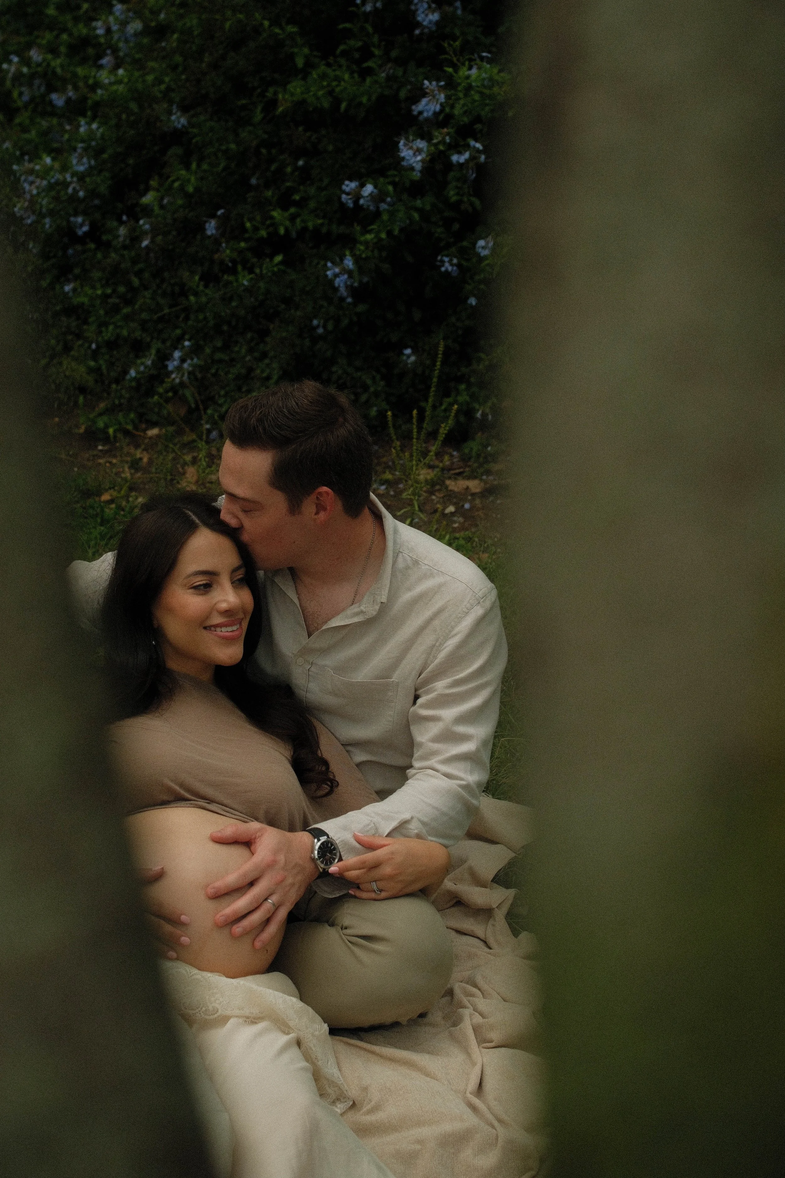 A couple sitting outdoors in front of a bush with small blue flowers, embracing each other with the woman resting her head on the man's shoulder and the man kissing her forehead.
