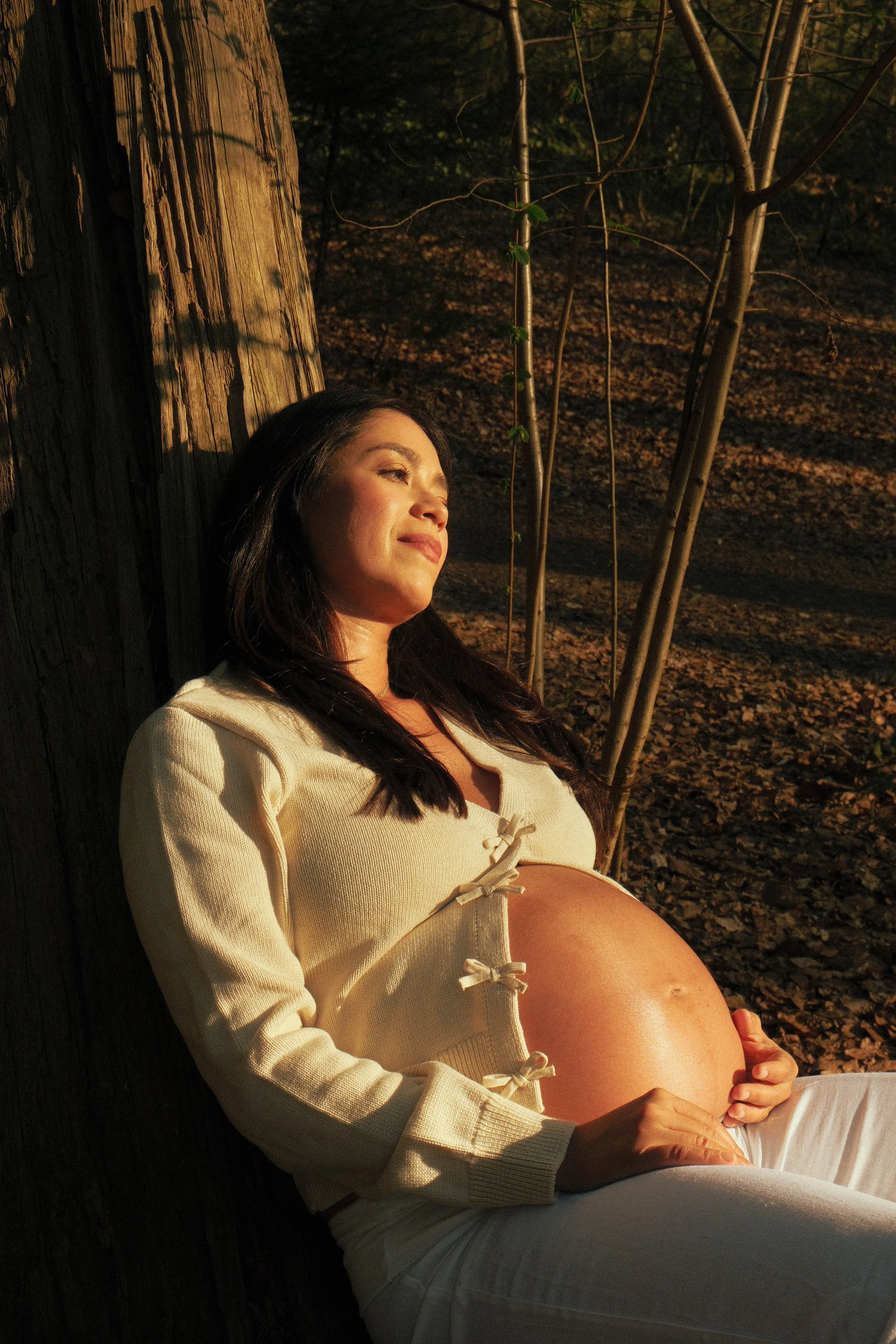 A pregnant woman with long dark hair relaxing outdoors against a tree trunk at sunset, wearing a cream-colored sweater with bows and white pants, with her hand resting on her belly.