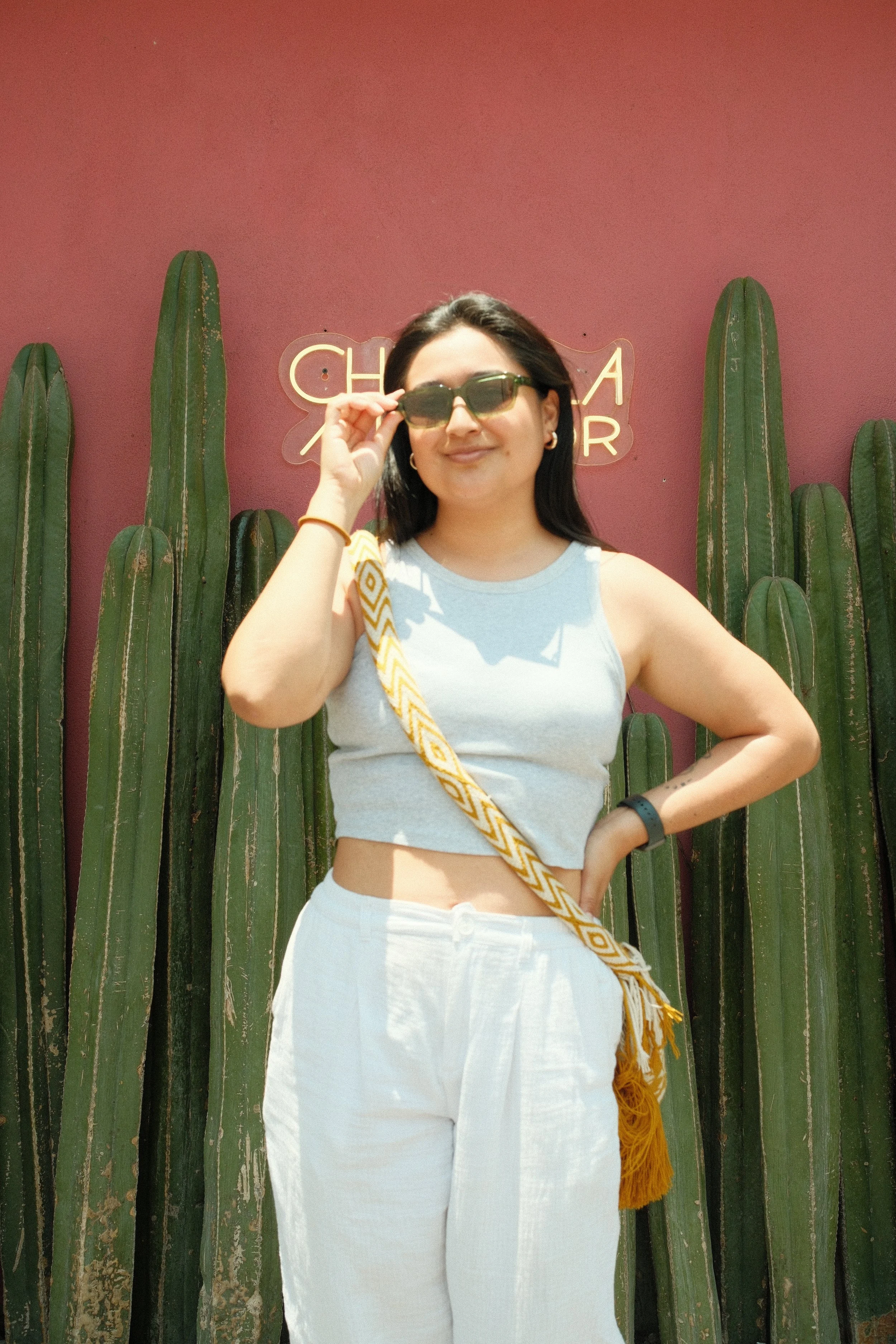 A woman wearing sunglasses, a white sleeveless top, and white pants standing in front of tall cactus plants on a pink wall with a neon sign.