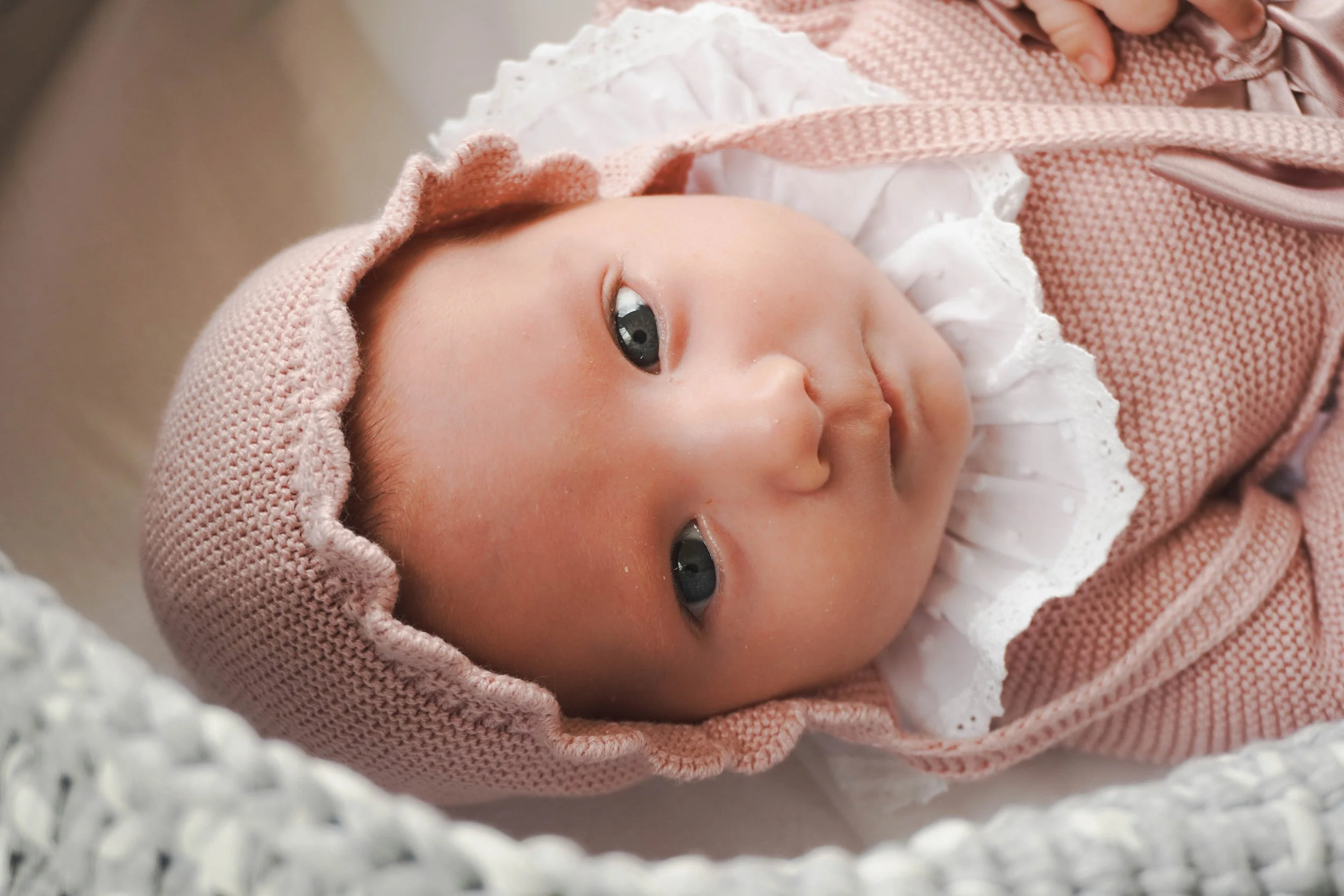 Close-up of a baby with blue eyes wearing a pink knit hat and pink outfit, lying on a white textured blanket.