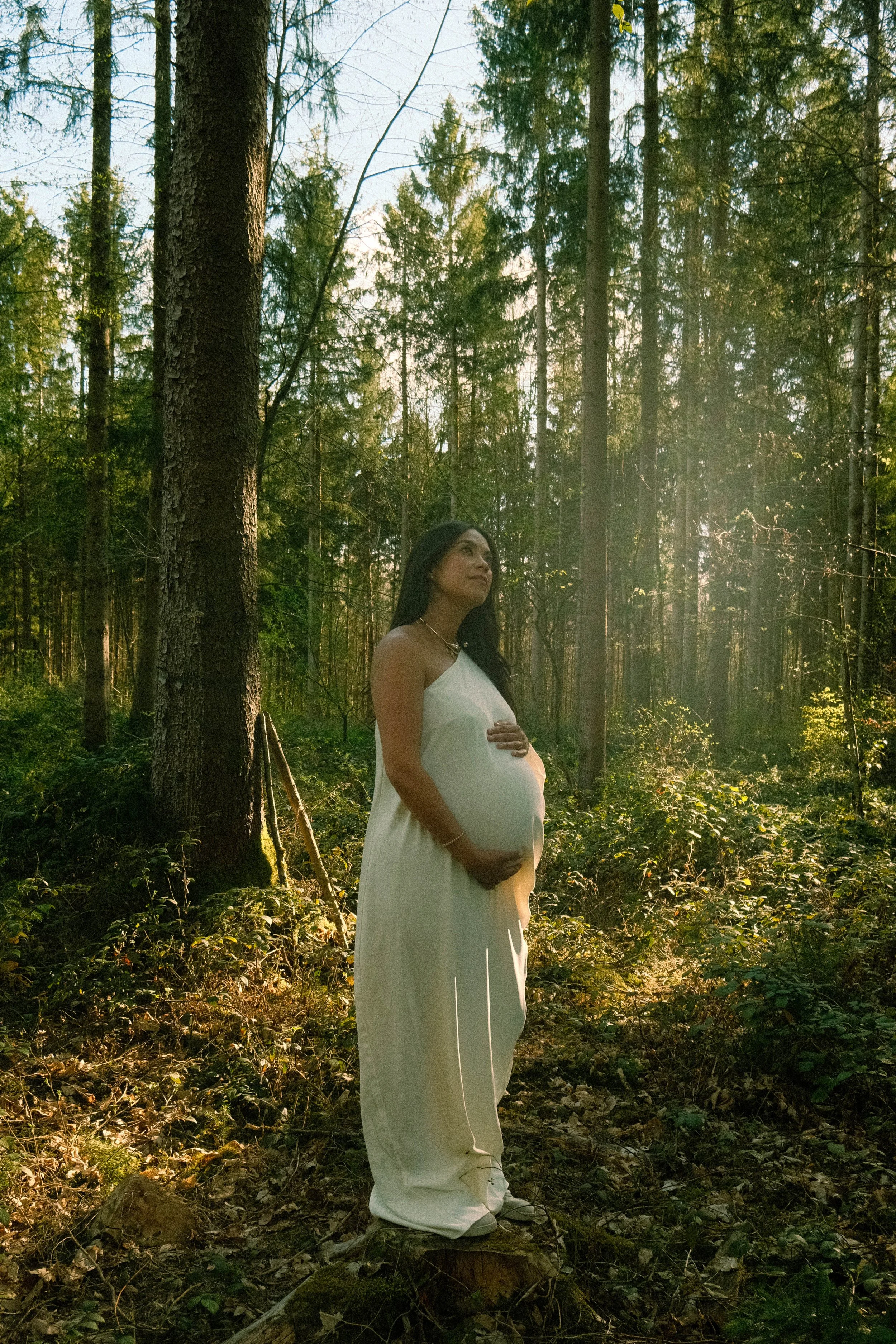 A pregnant woman in a white dress standing in a sunlit forest holding her belly with both hands.