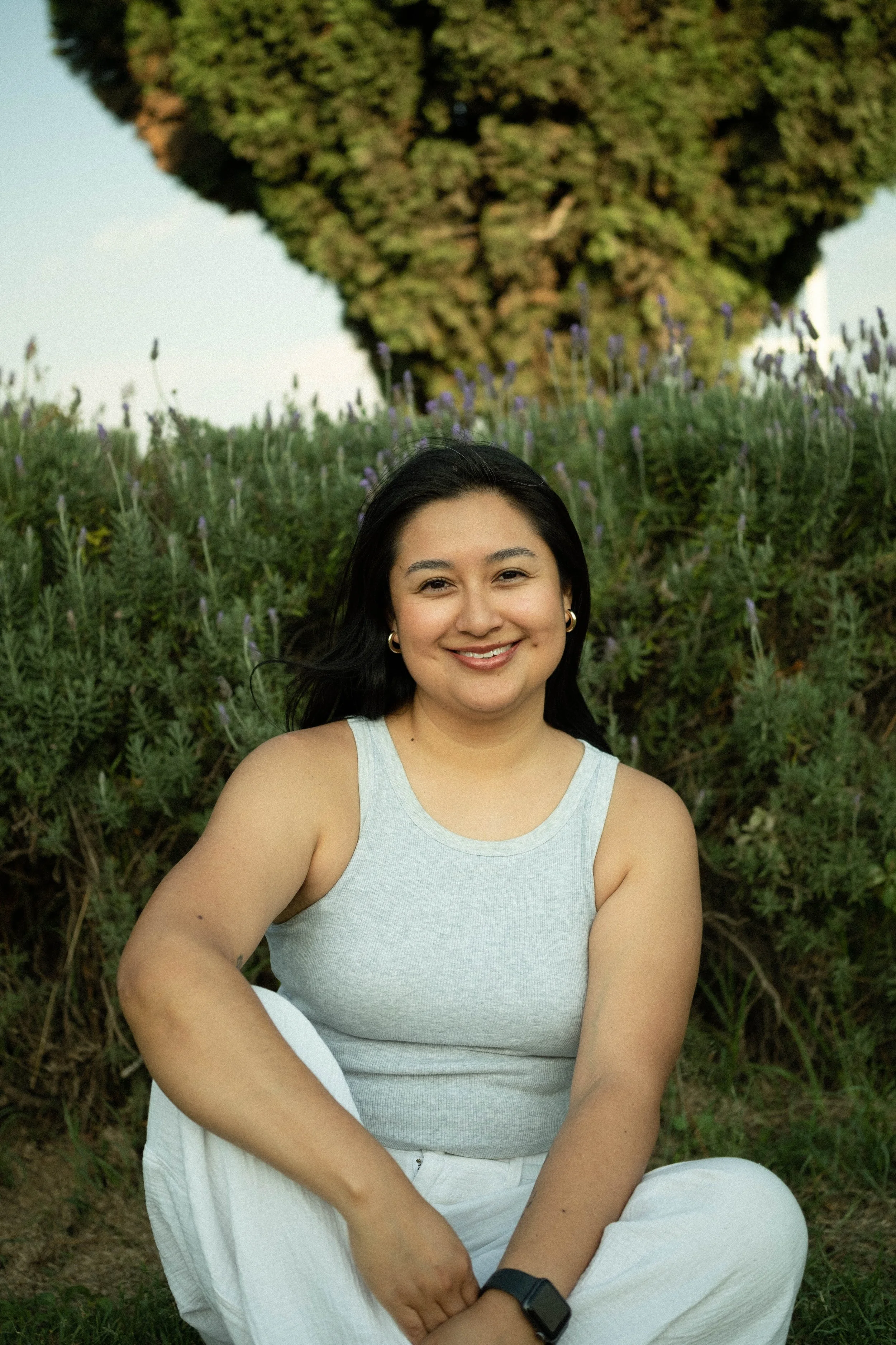 A smiling woman with long dark hair, wearing a gray tank top and white pants, sitting outdoors on the grass with purple flowers and a large tree in the background.