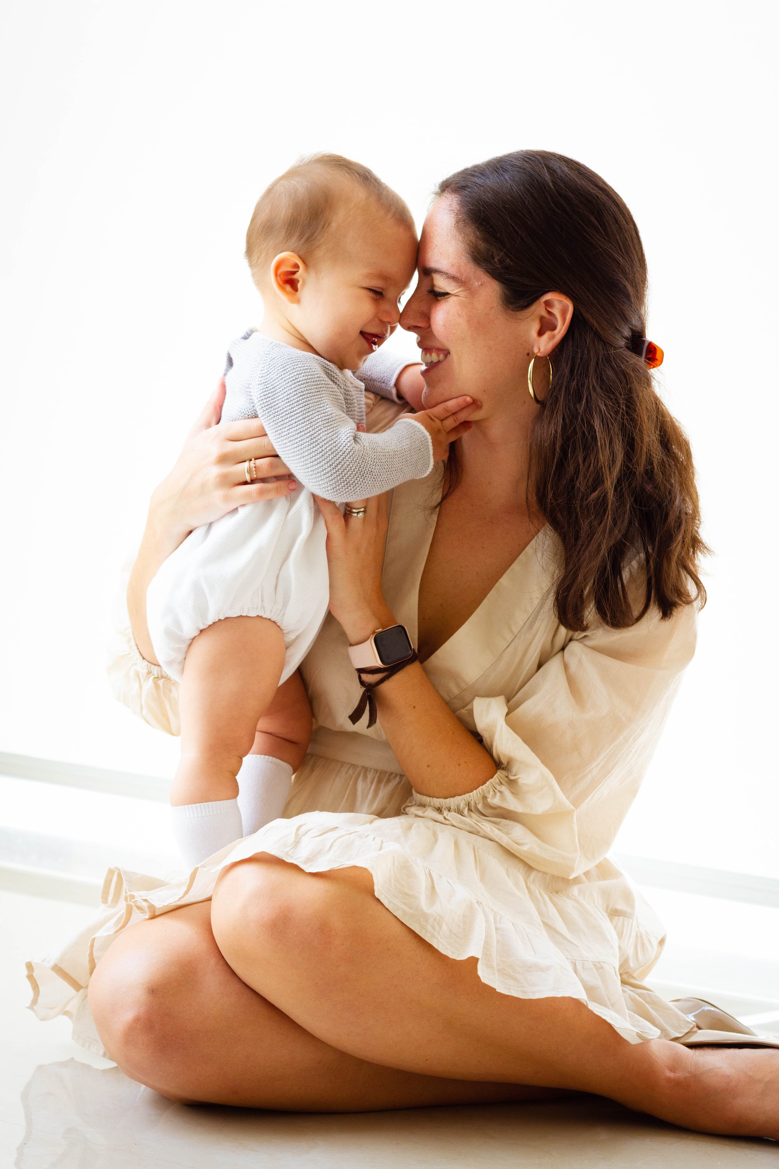 A woman with brown hair, wearing earrings and a beige dress, is sitting on the floor with her legs crossed, holding a laughing young boy with short hair, wearing a gray sweater and white shorts, close to her face, both touching their foreheads togeth