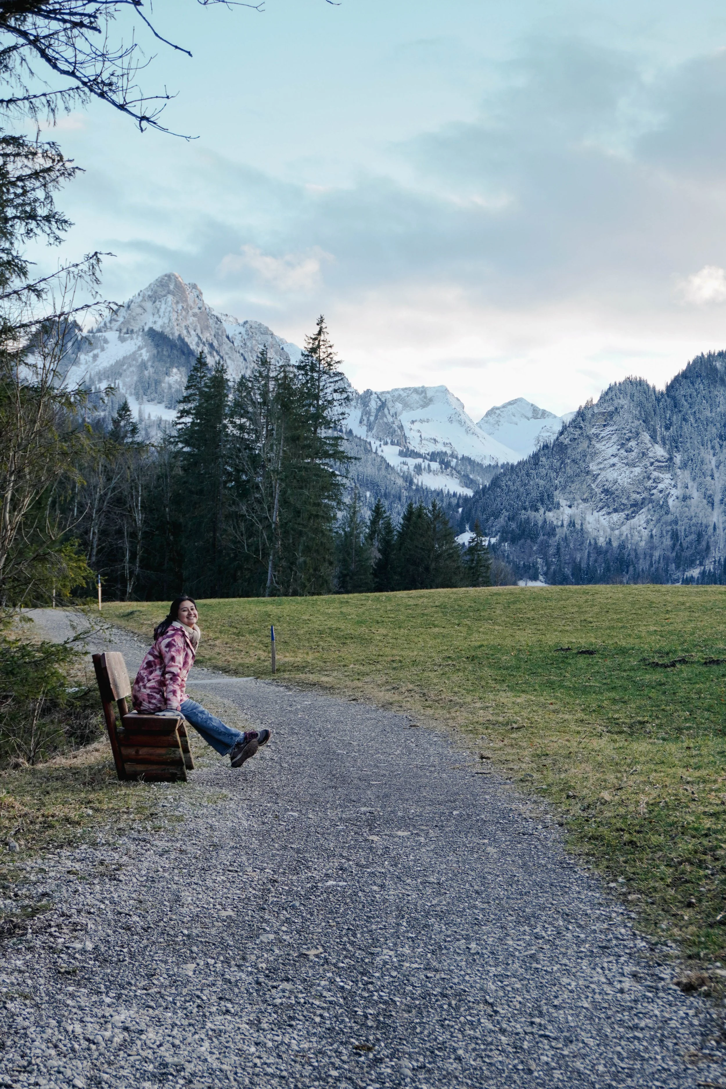 A woman sitting on a wooden bench on a gravel path in a park with snow-capped mountains and evergreen trees in the background, partly cloudy sky.