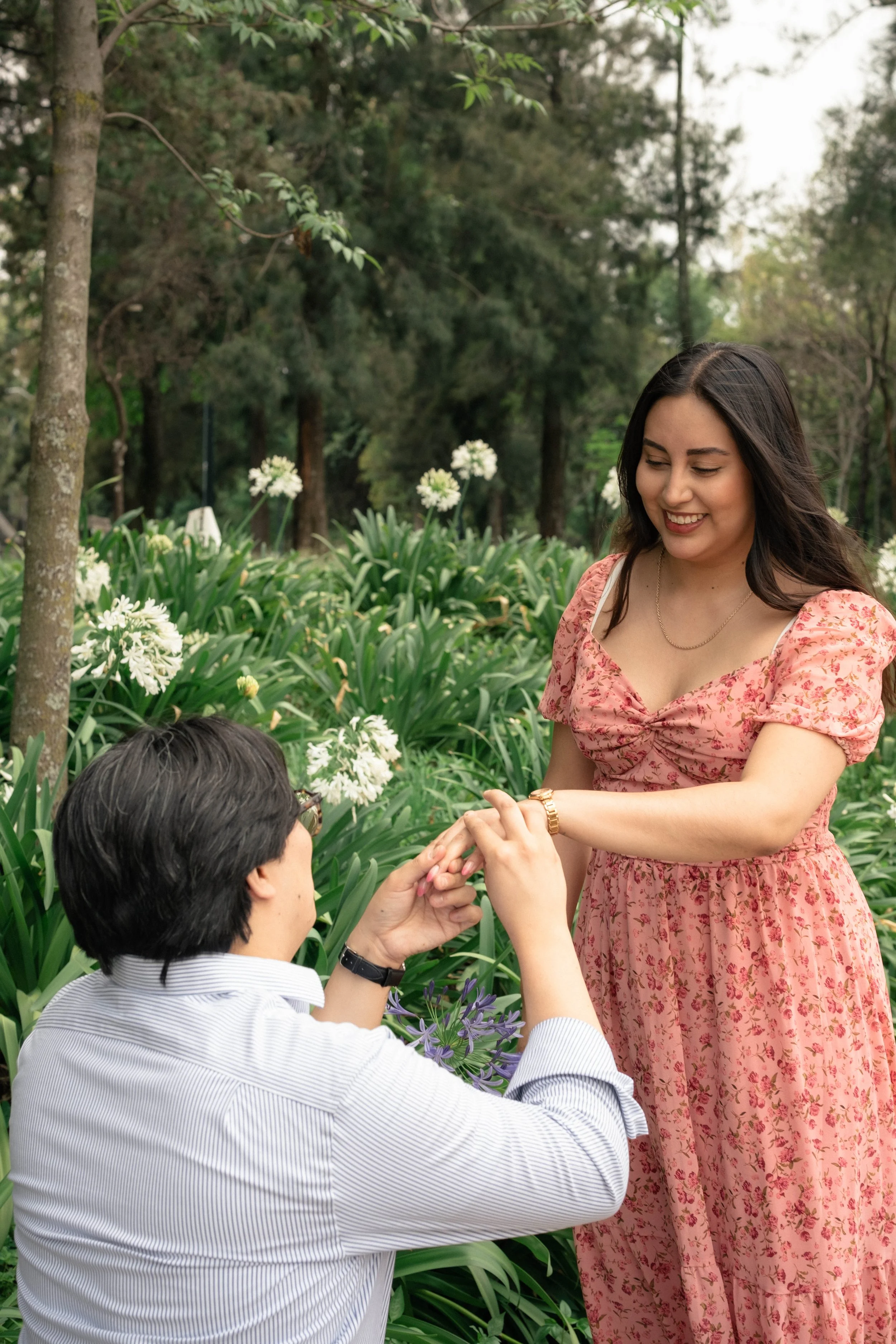 A man kneels in a garden with purple flowers, proposing marriage by holding a woman's hands. The woman, standing, has dark hair, is smiling, and wears a pink floral dress.