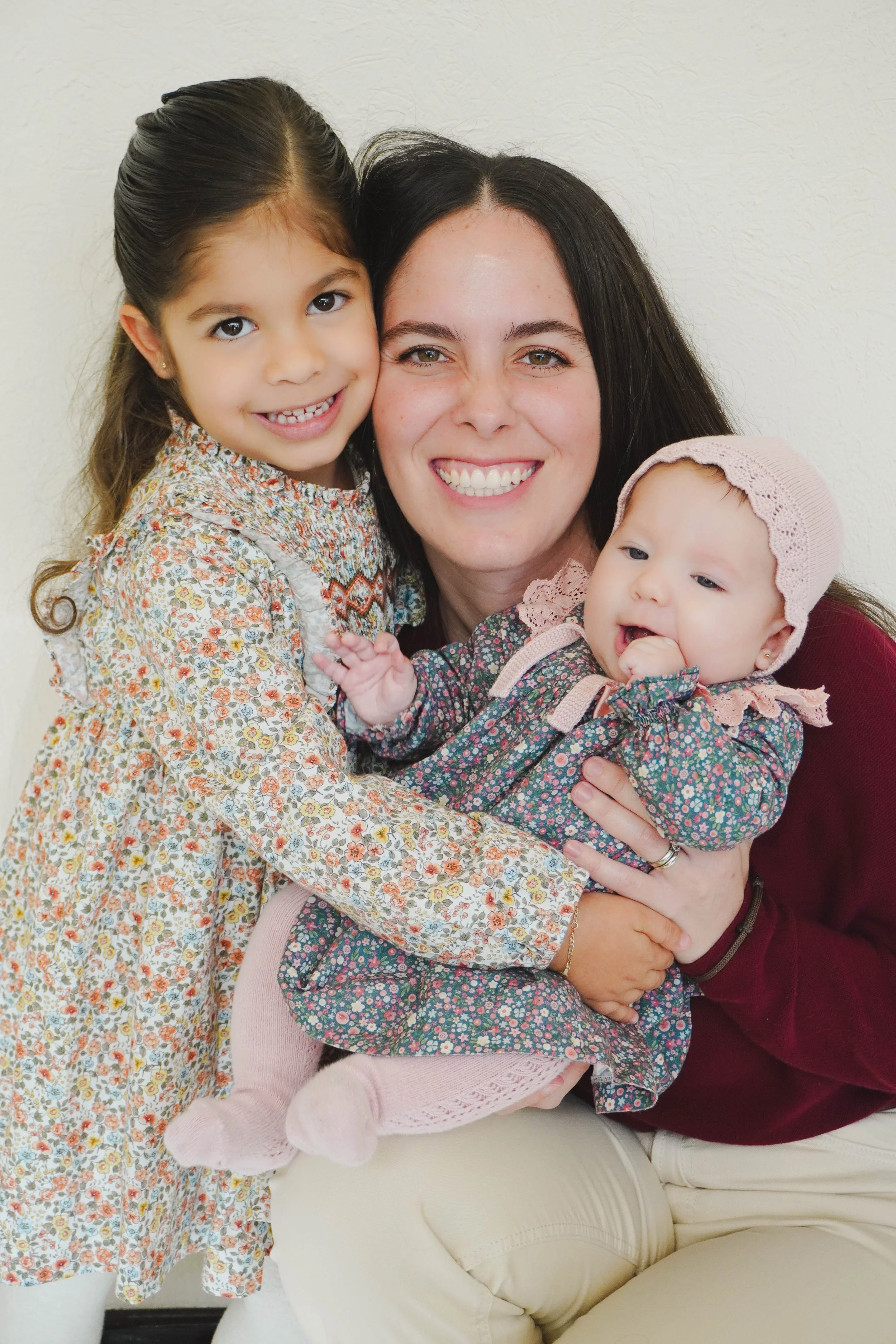 A woman smiling with a young girl and a baby girl in her arms in front of a plain white wall.
