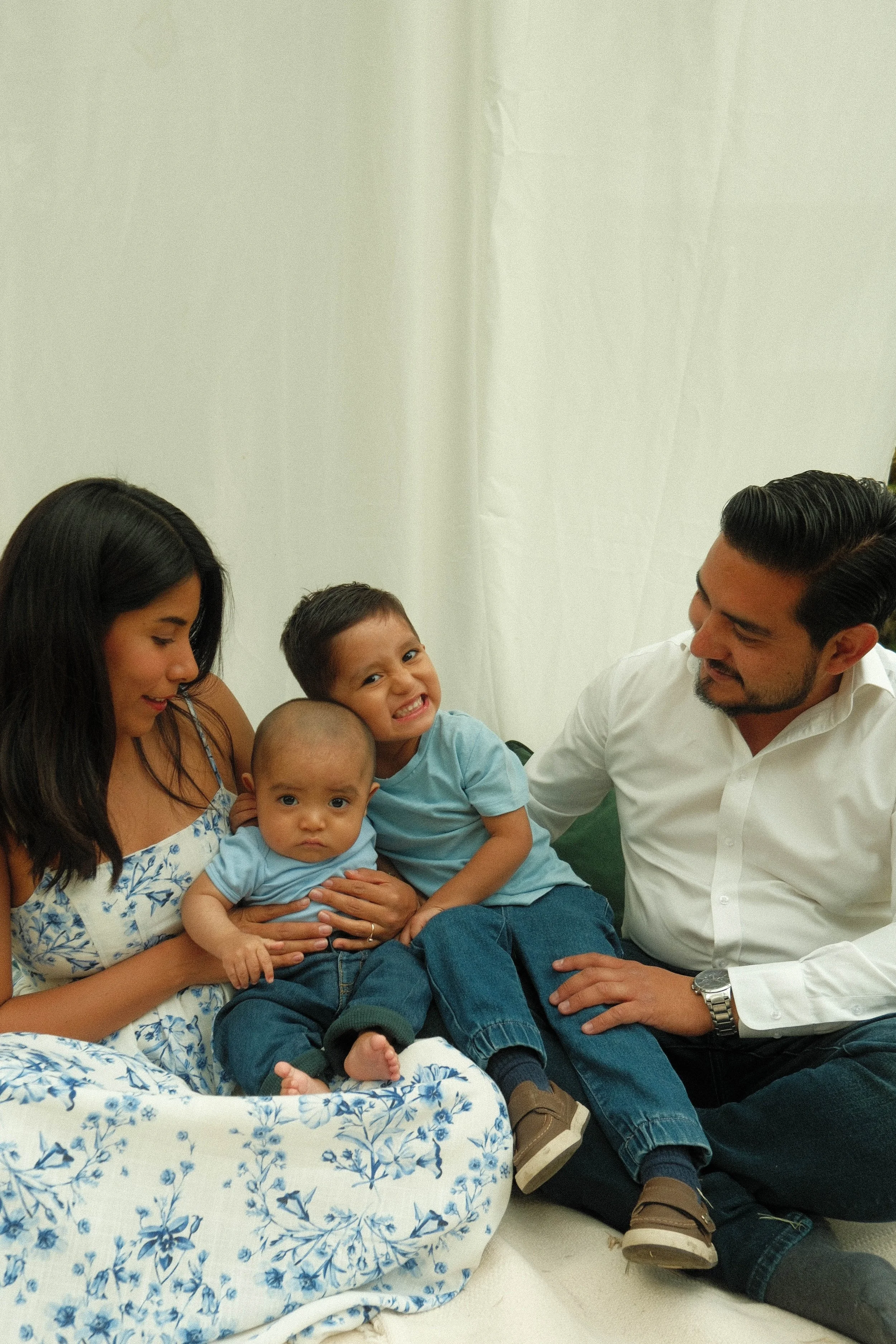 A family group sits together, including two adults and three children, with the children sitting on a woman’s lap. They are in a bright indoor setting, with a mostly plain backdrop. The children are young, and the woman appears to be pregnant. All ar