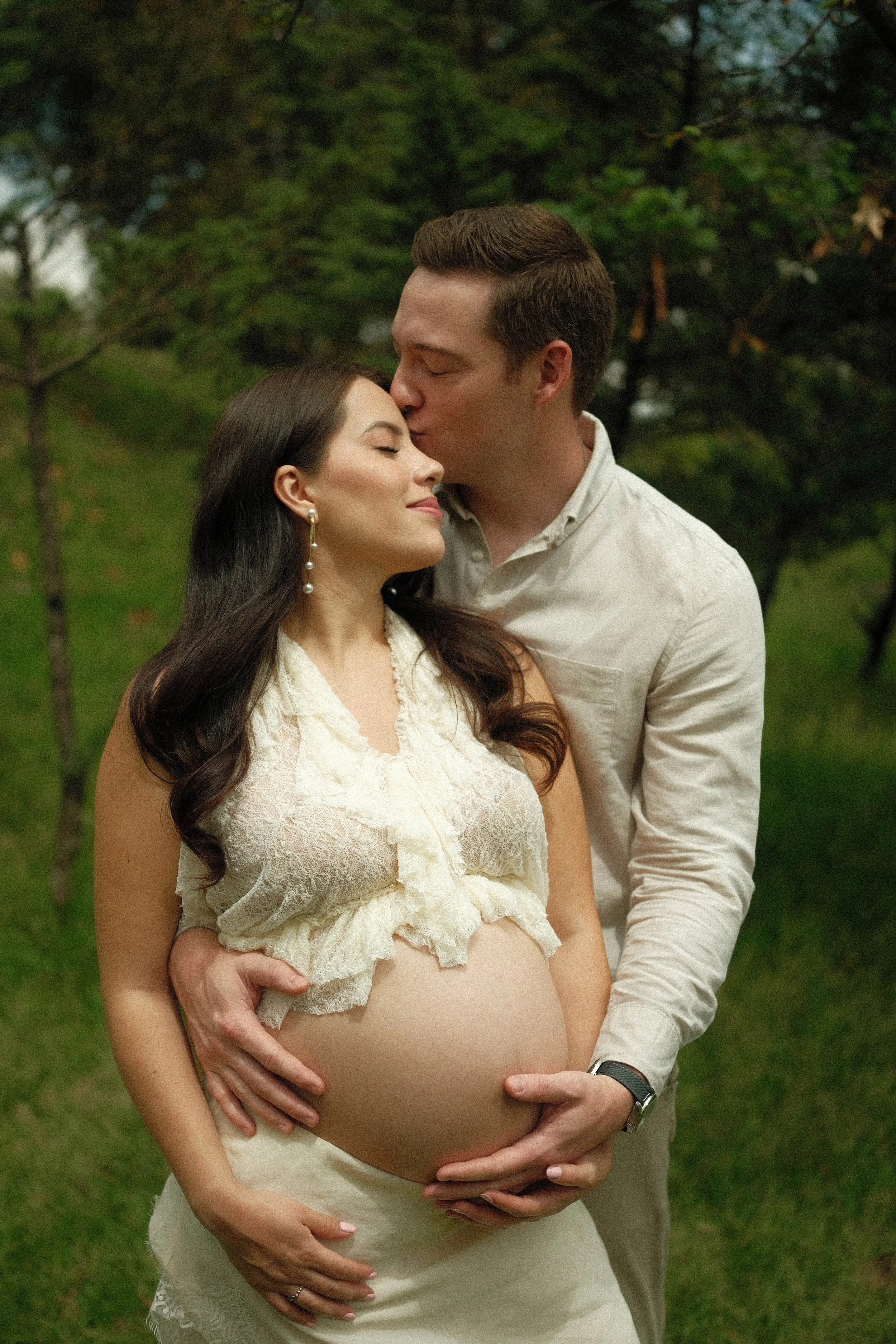 A pregnant woman in a cream lace dress being embraced by a man in a light-colored shirt, holding her belly and giving her a kiss on the forehead in a green outdoor setting.