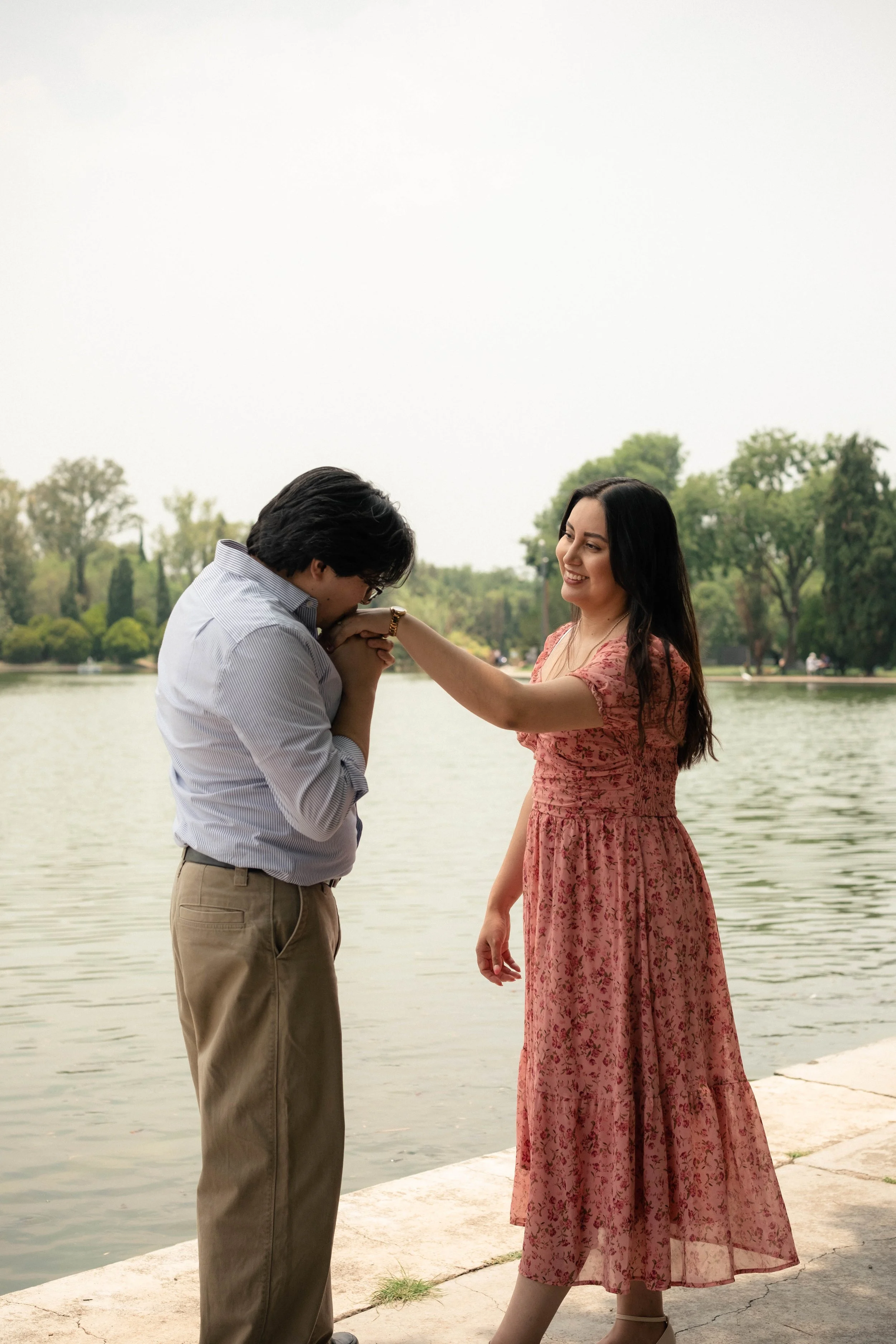 A man kneeling and holding a woman's hand by a lake. The woman is smiling and wearing a pink floral dress. The man is wearing a striped shirt and khaki pants. Trees are in the background.