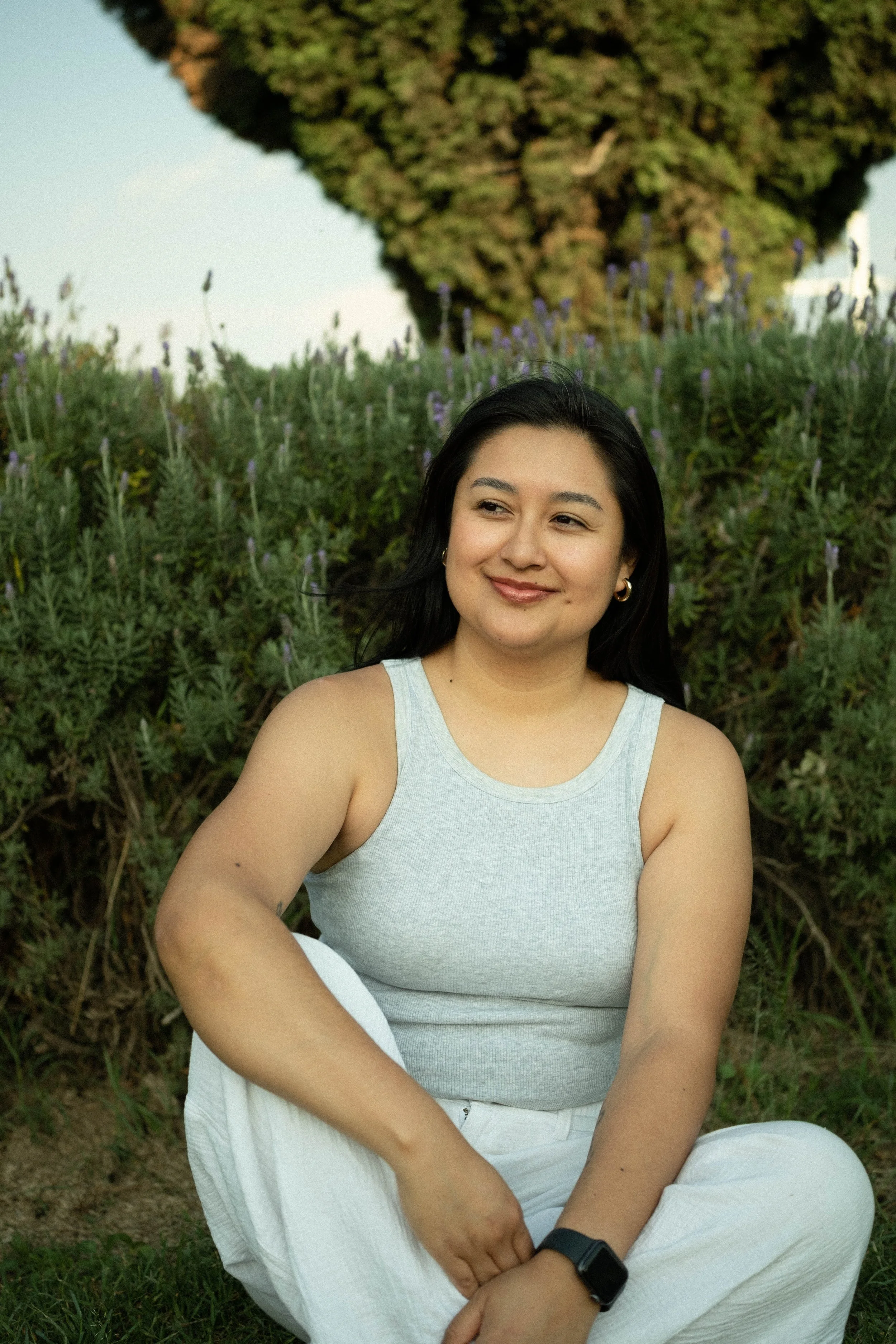 Young woman with dark hair smiling, sitting outdoors in front of greenery and purple flowers, wearing a light gray tank top, white pants, and a black smartwatch.