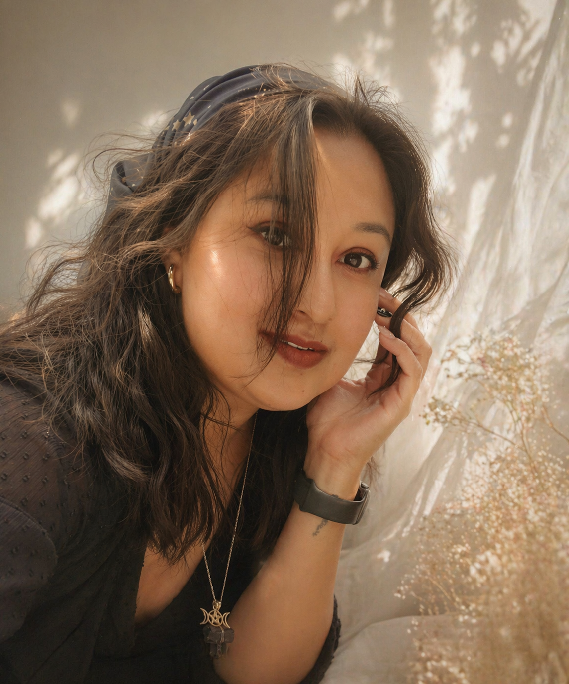 A woman with wavy brown hair, wearing a black headscarf, gold earrings, and a necklace, smiling softly, resting her face on her hand, with a blurred background of trees and sunlight.