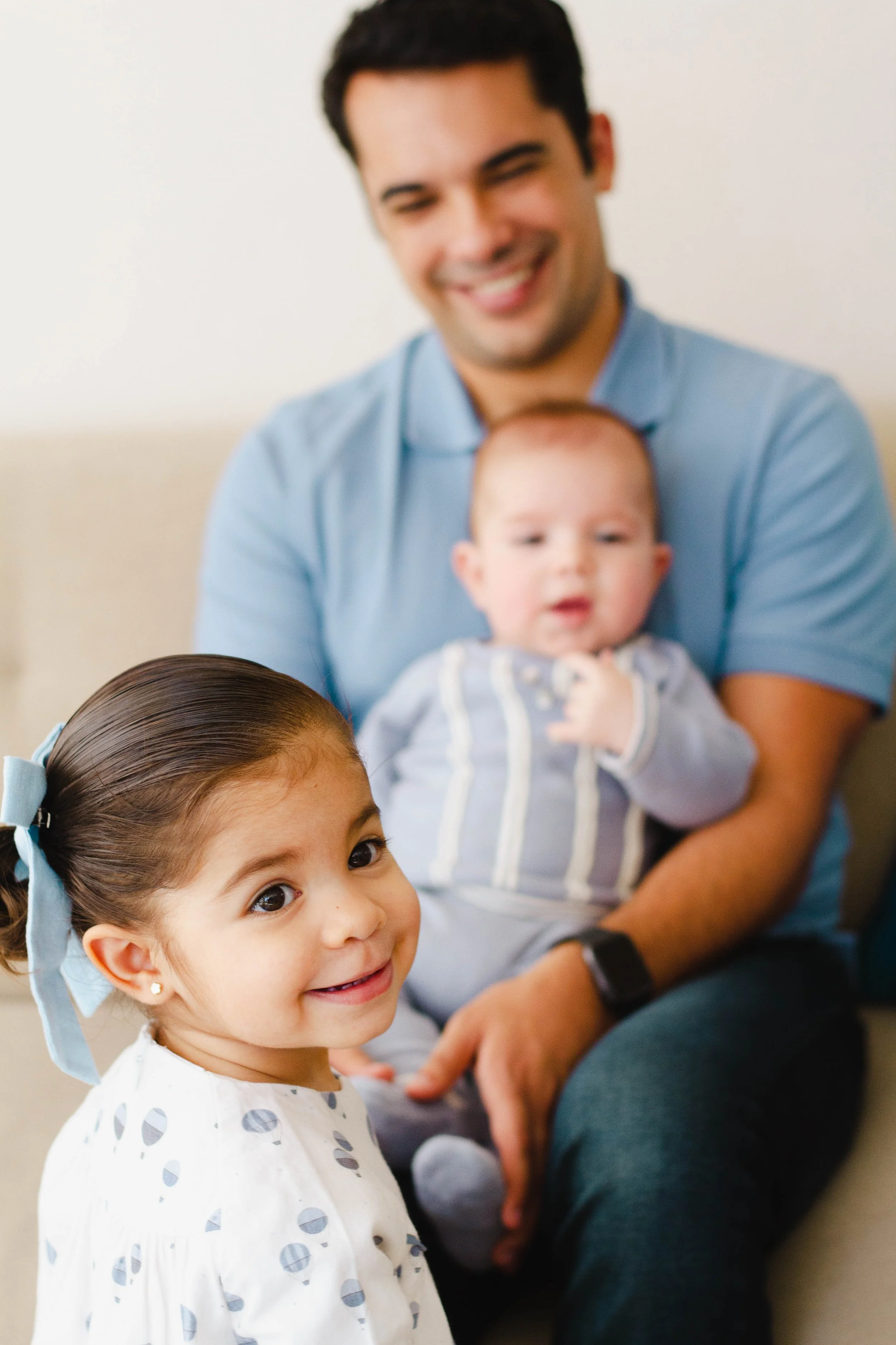 A young girl with brown hair, wearing a white shirt with grey polka dots, smiling and looking at the camera. In the background, a man with dark hair wearing a blue shirt is holding a baby, who is blurry, and smiling with big eyes.