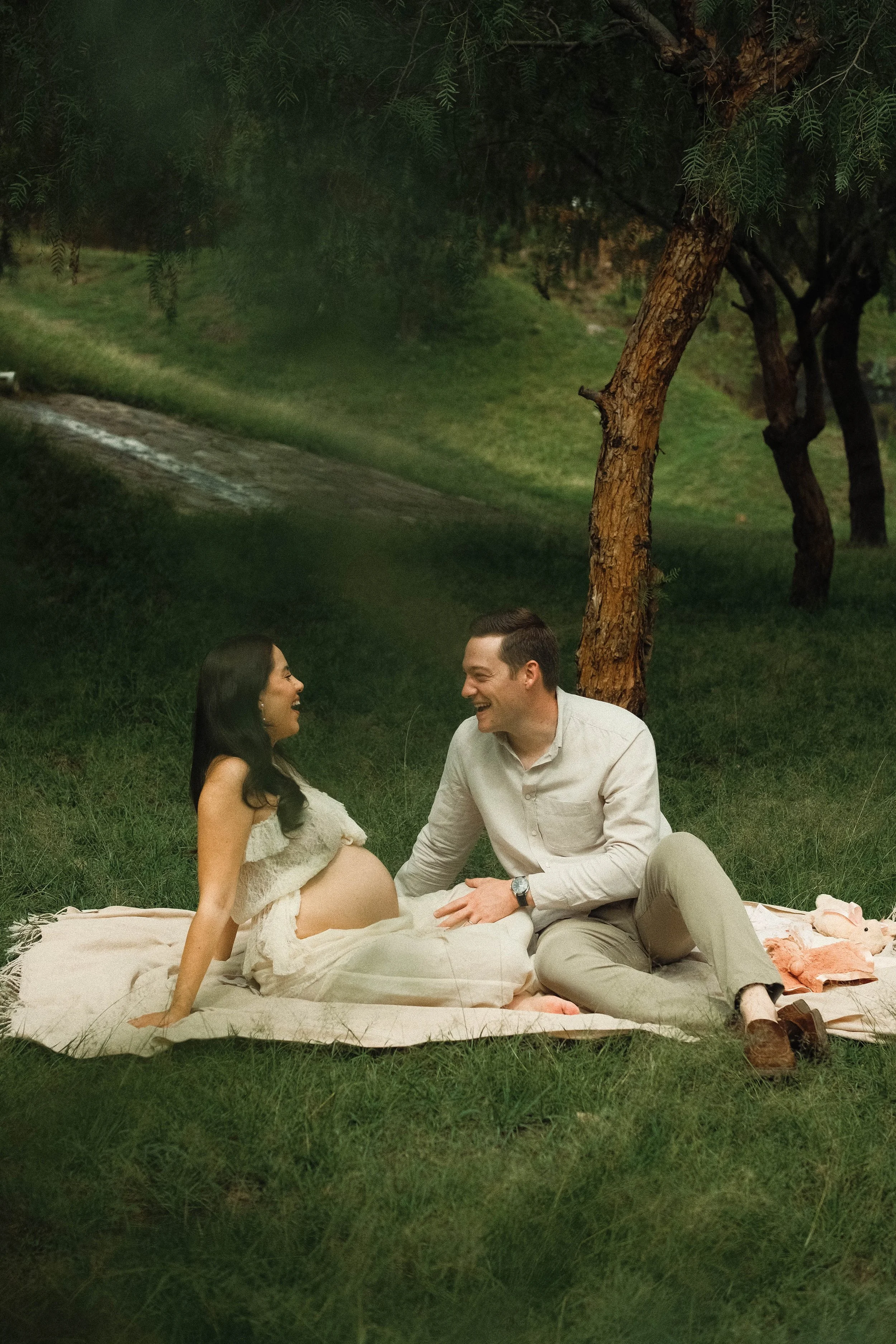 A pregnant woman and a man sitting outdoors on a blanket, smiling at each other, in a park at night. The woman is wearing a light dress and the man a light shirt and pants. Trees and grass surround them.