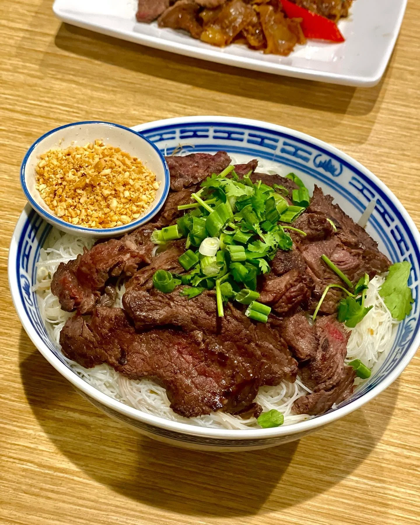 Bowl of cooked beef slices on rice noodles topped with chopped green onions, served with crushed peanuts in a small dish, on a wooden table.