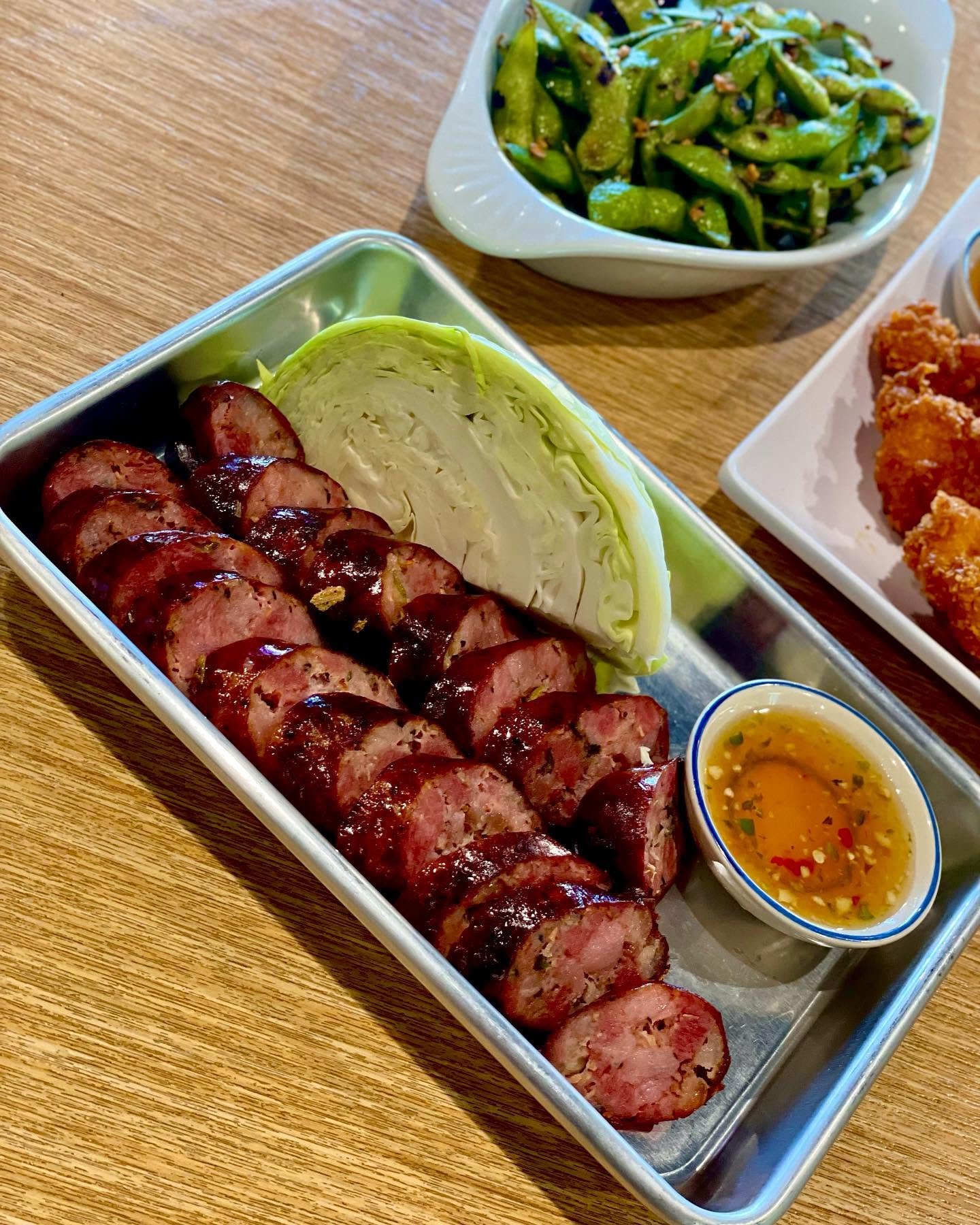 Sliced sausage with ketchup on a metal tray, sliced cabbage, bowl of dipping sauce, bowl of green beans, fried chicken pieces
