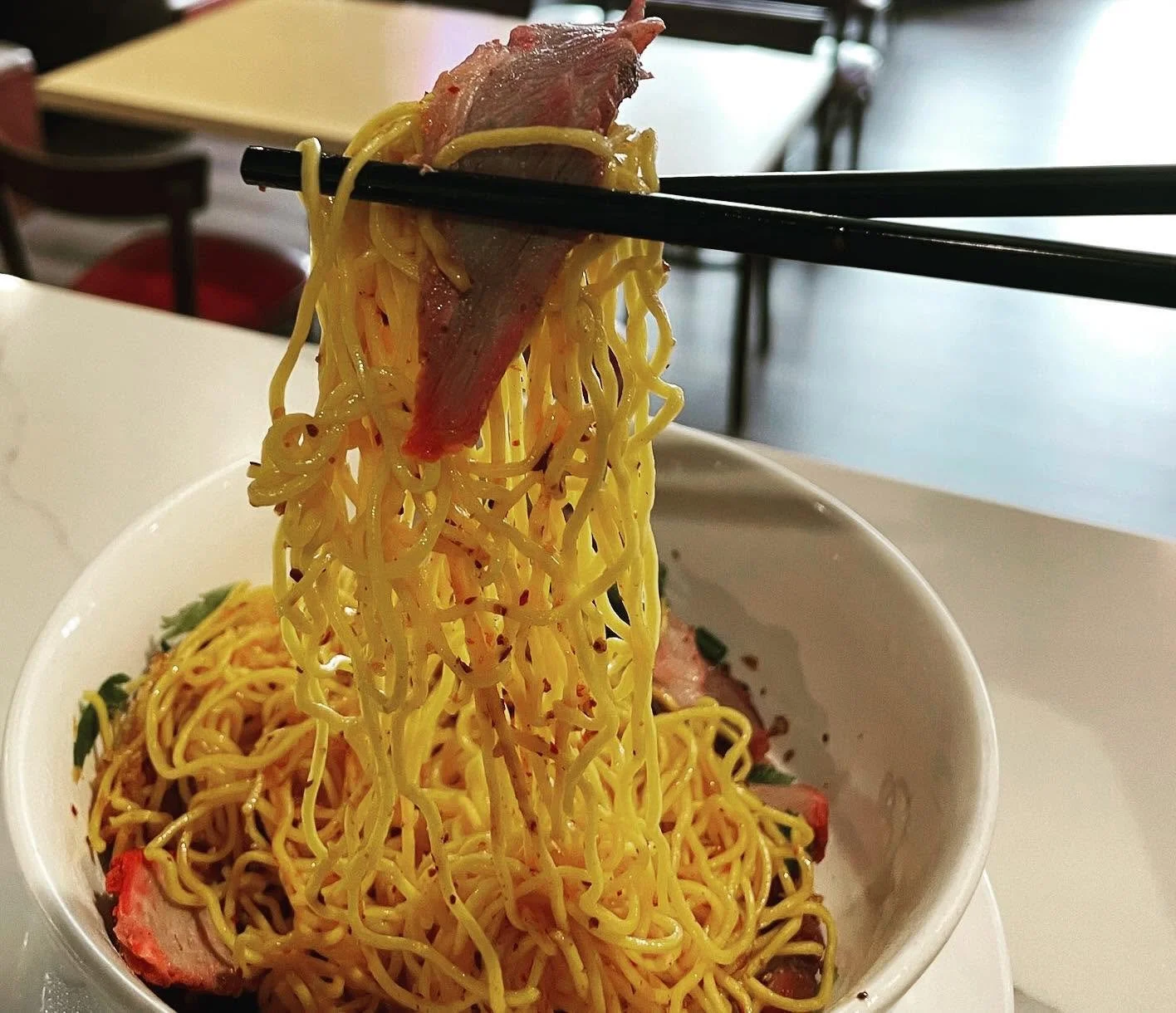 Chopsticks lifting a portion of cooked yellow ramen noodles with slices of meat in a white bowl.