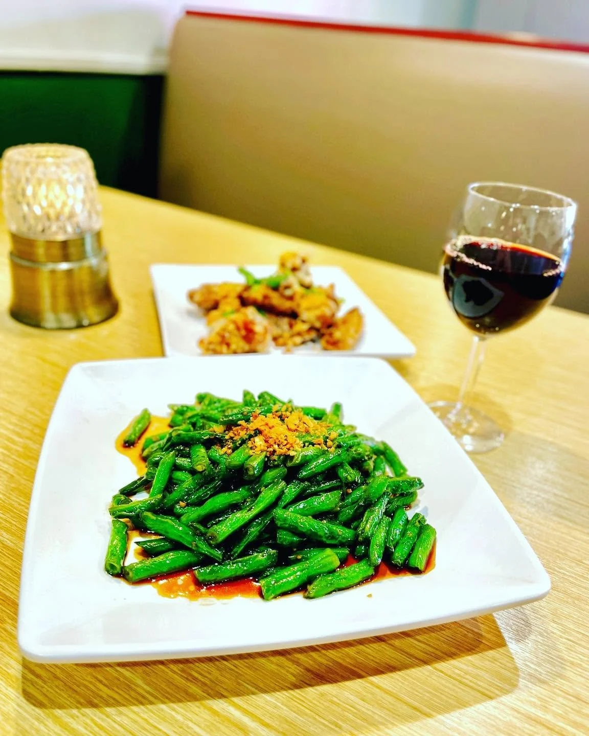 A table with two white square plates of food, a glass of red wine, and a candle. The front plate has green beans with a crumb topping and sauce. The back plate has fried food garnished with green herbs.