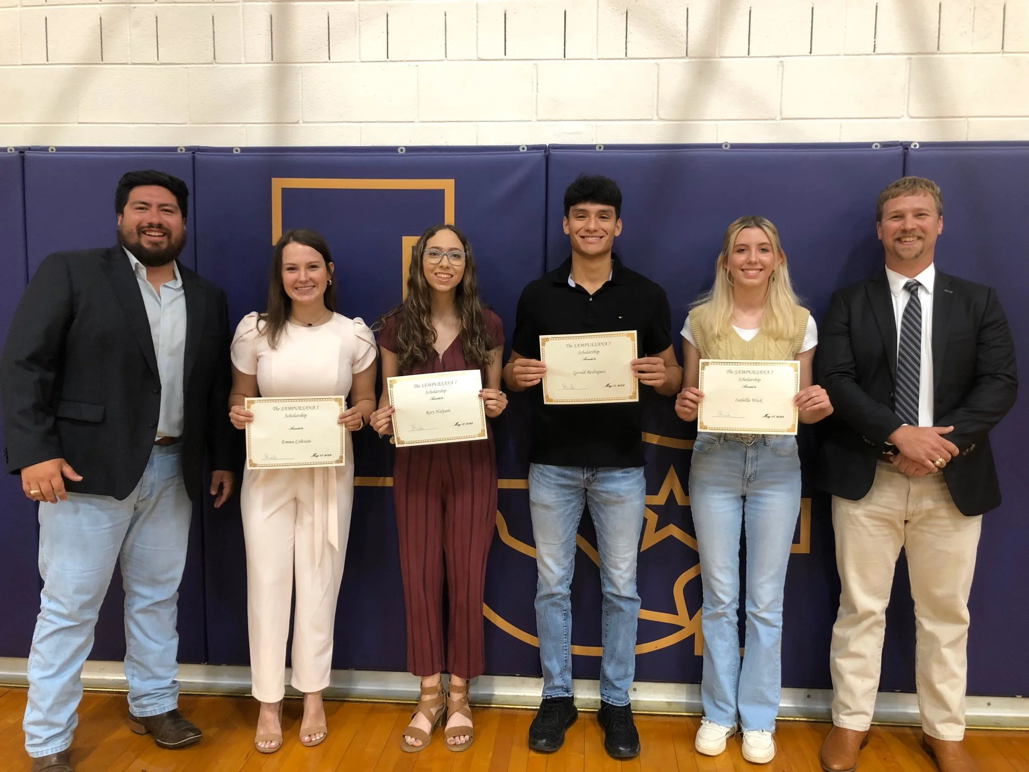 Five people, three men and two women, standing in a line holding certificates, in front of a blue and gold wall with a star emblem, smiling for a photo.
