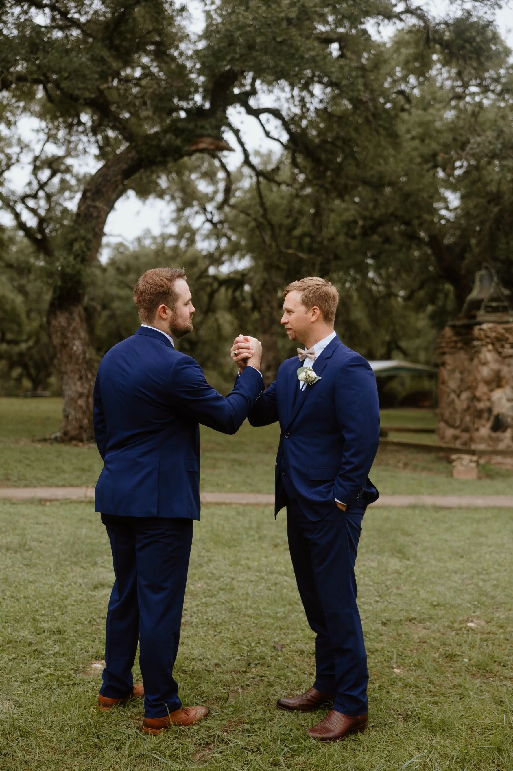 Two men in tuxedos shaking hands outdoors in a park with large trees in the background, one with a boutonniere, indicating a wedding or formal event.