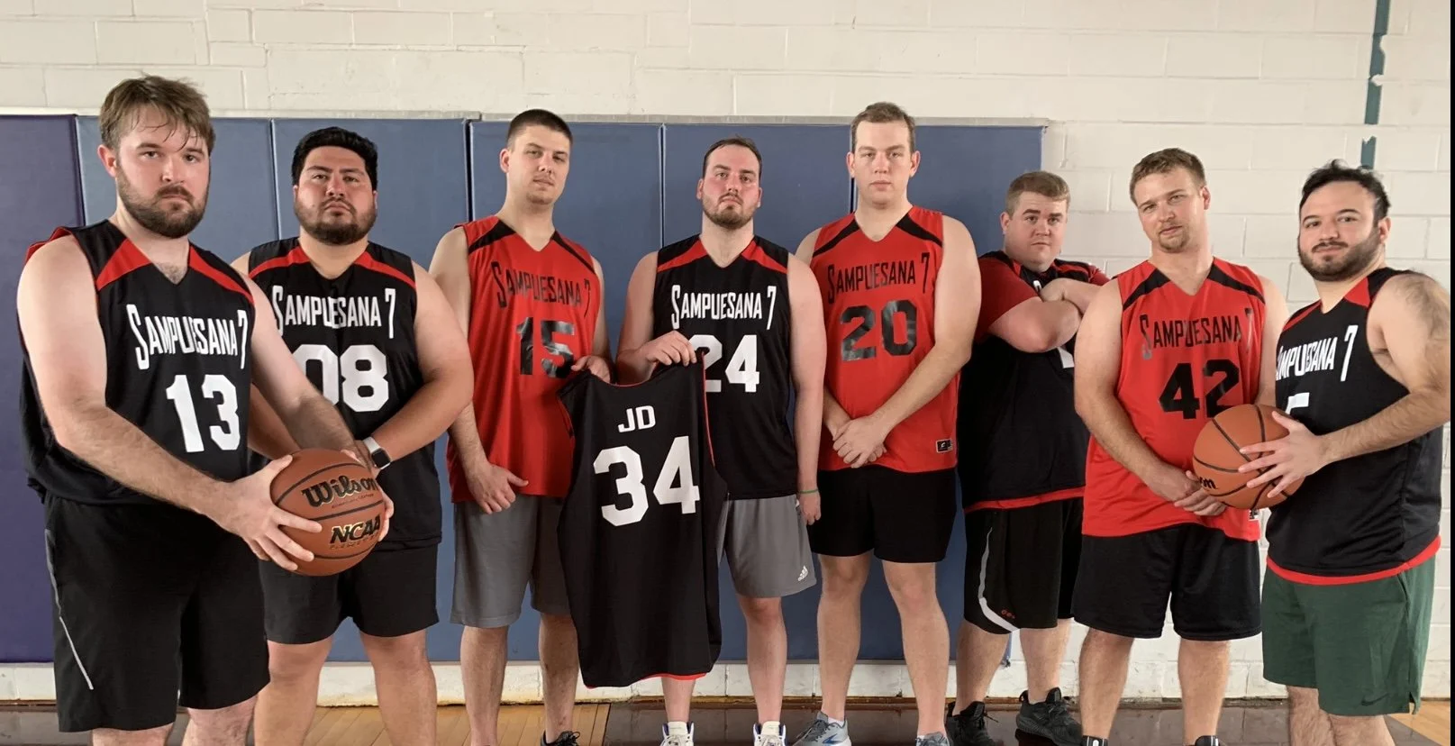 Group of men in basketball jerseys standing in a gymnasium, some holding basketballs.