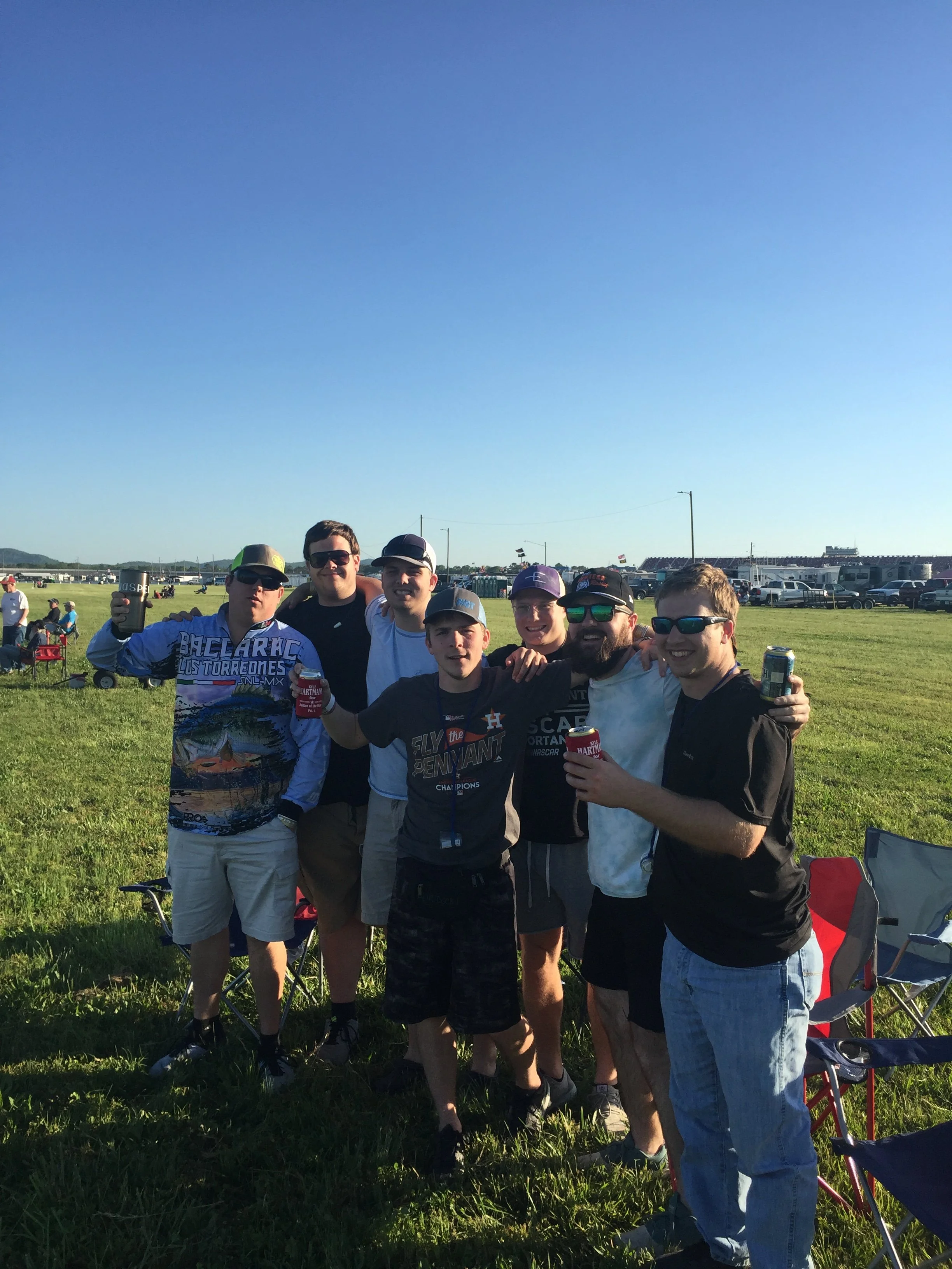 Group of friends standing outdoors on a sunny day, smiling and posing for a photo at a race track or sporting event, with cars and tents in the background.