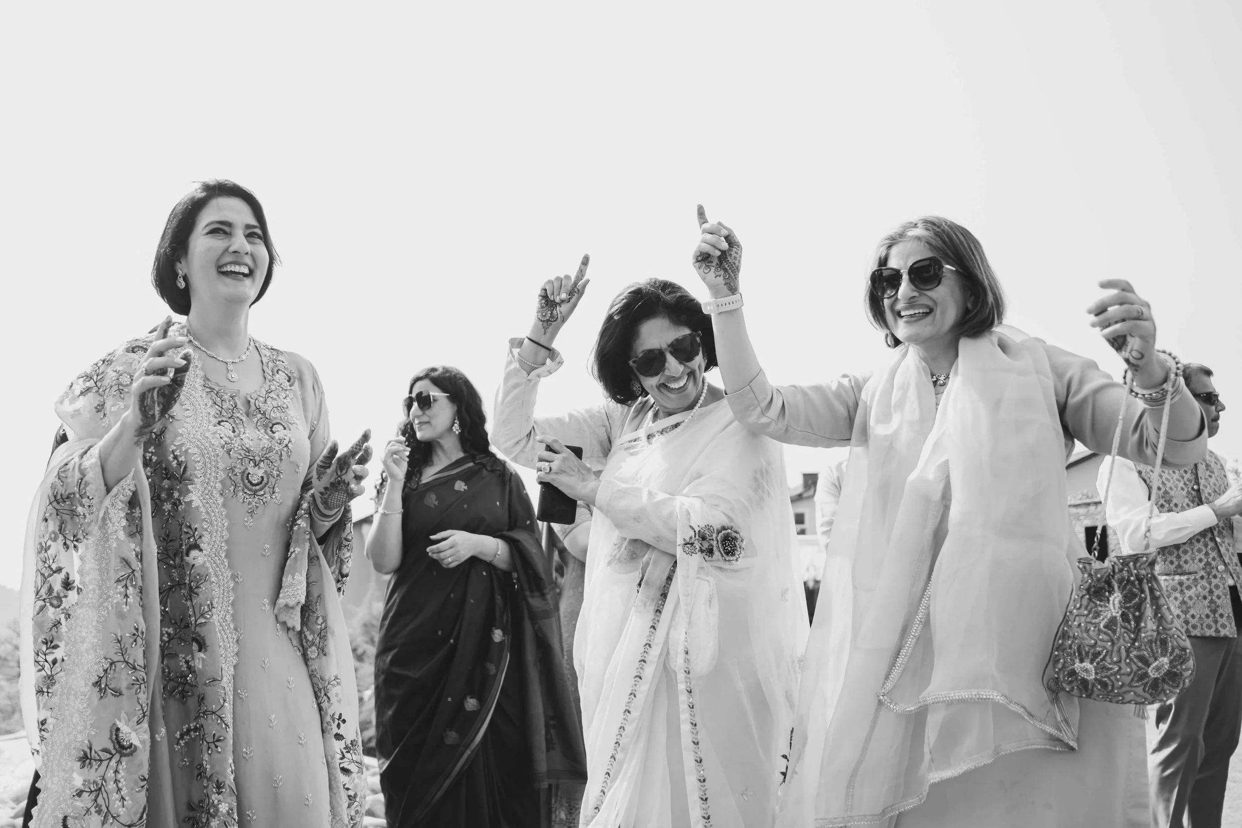 Group of women smiling and dancing outdoors, dressed in traditional South Asian attire, some wearing sunglasses, with a clear sky in the background.
