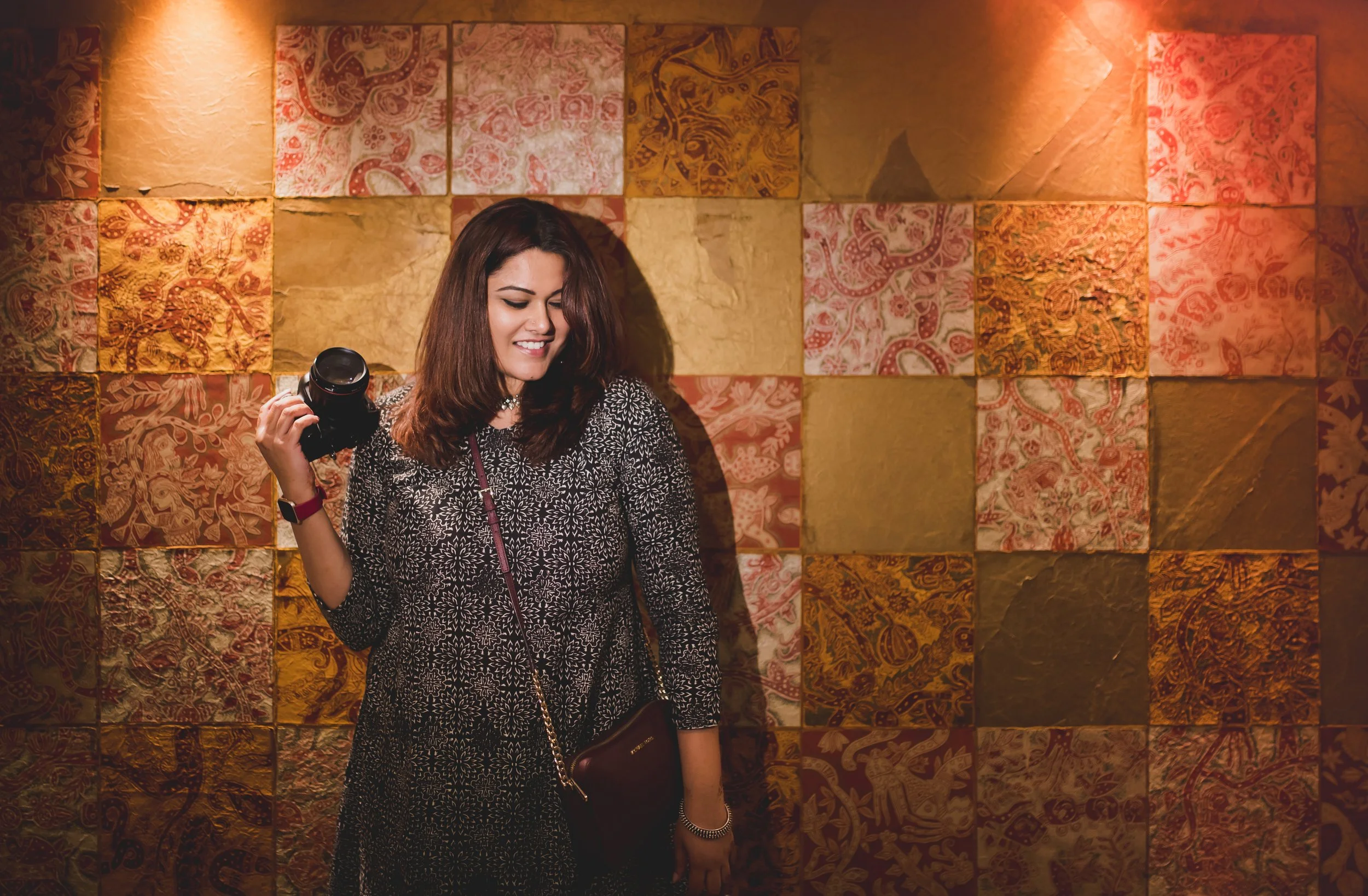 A woman with brown hair holding a camera, standing in front of a colorful wall with patterned tiles.