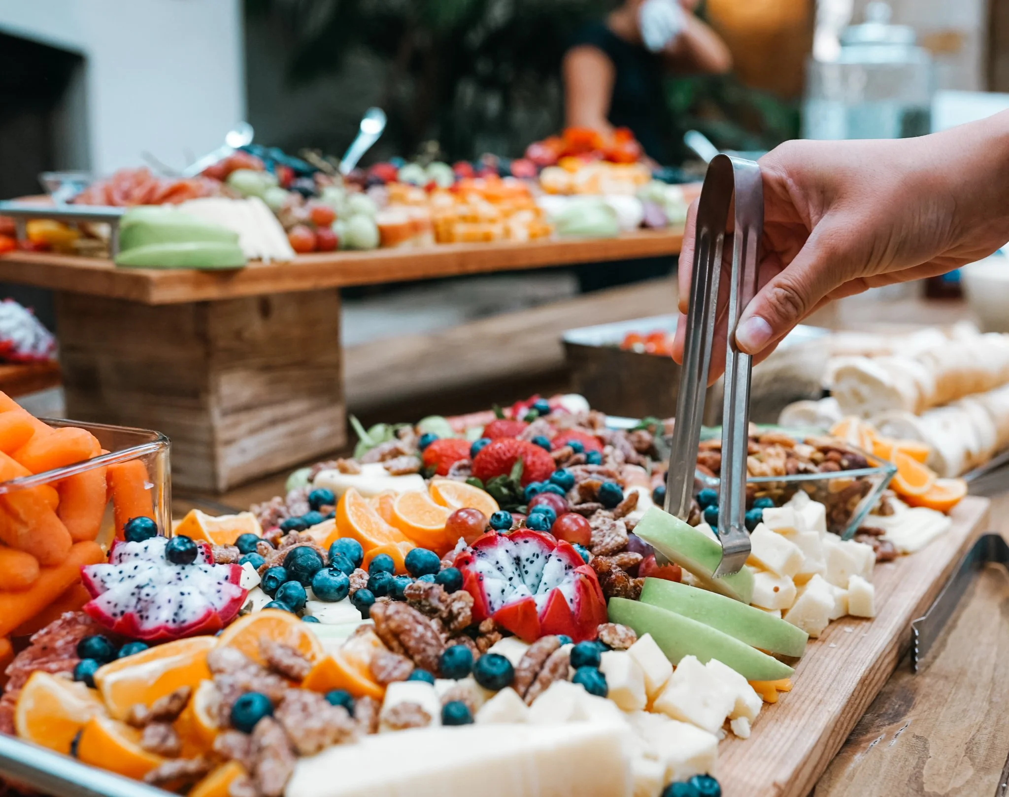 A person using tongs to serve themselves fruit and cheese on a platter at a buffet or party. The platter contains various berries, sliced oranges, and pieces of cheese.